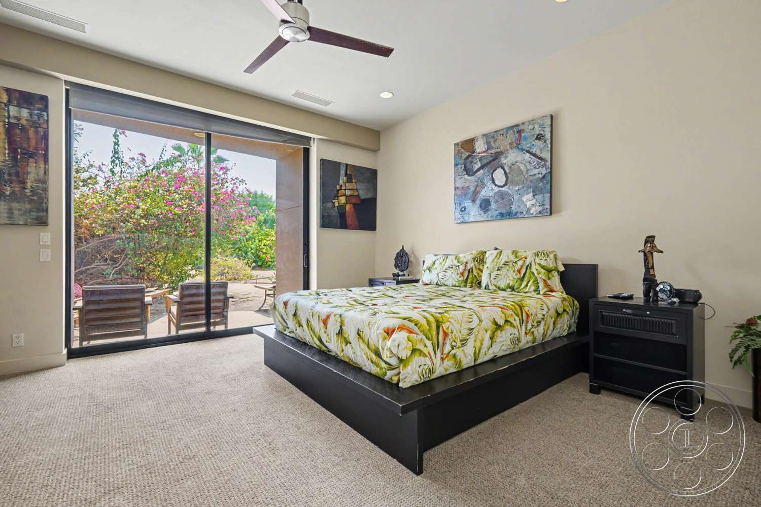 Rancho Mirage 4 - ceiling fan, floor-to-ceiling windows, decorative wall art, black nightstand, neutral painted walls, modern bed frame, carpet bedroom flooring, sliding glass patio door, outdoor seating area view, floral bedding pattern, natural light lighting