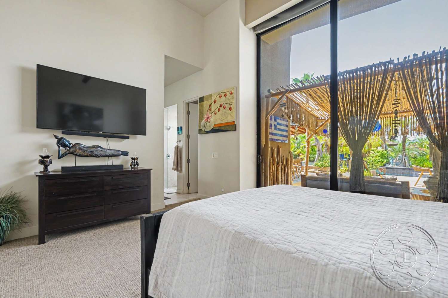 Rancho Mirage 4 - natural light, floor-to-ceiling windows, textured wall treatment, dark wood dresser, outdoor patio view, framed artwork on wall, carpet bedroom flooring, open doorway to bathroom, large flat-screen TV wall-mounted, modern indoor plants, luxe bedding