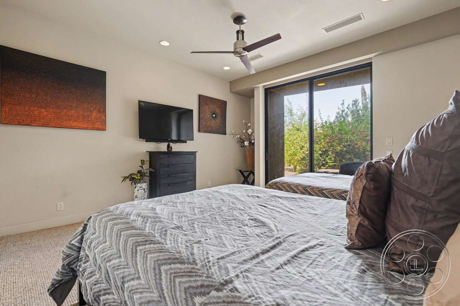 Rancho Mirage 4 - neutral color palette, wooden dresser, ceiling fan, floor-to-ceiling windows, decorative wall art, textured plaster walls, beige bedding, flat-screen television, carpet bedroom flooring, black wood nightstand, green outdoor view