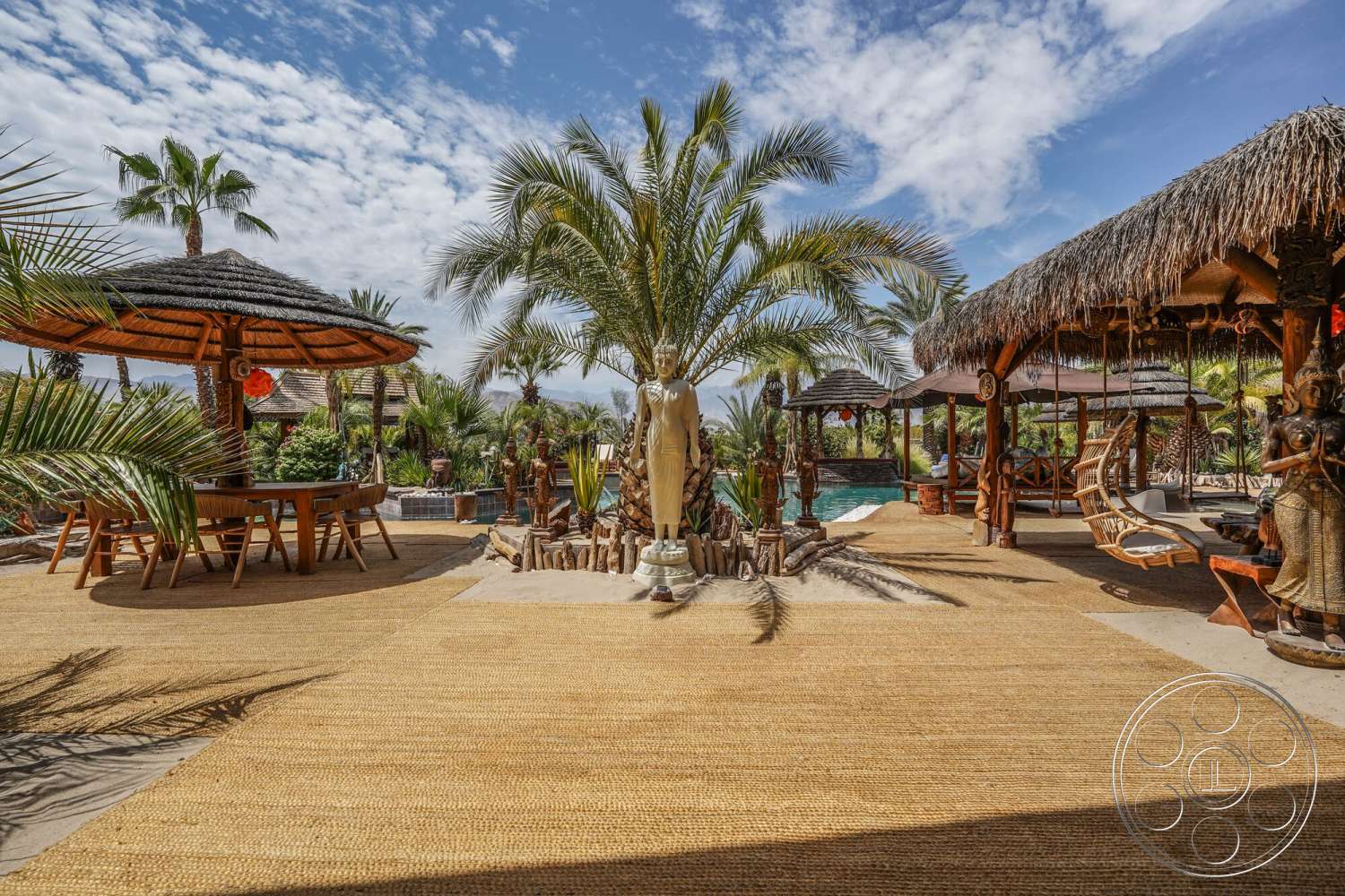 Rancho Mirage 4 - exterior, thatched roof gazebo, ceramic planters, outdoor lounging area, tropical palm trees, sunny outdoor setting, lush garden landscaping, wooden outdoor dining table, woven grass patio floor, stone sculpture garden, wooden swing chair, decorative wooden carvings, natural stone pool edge, poolside with reflection, rustic wooden pillars, tropical themed paradise