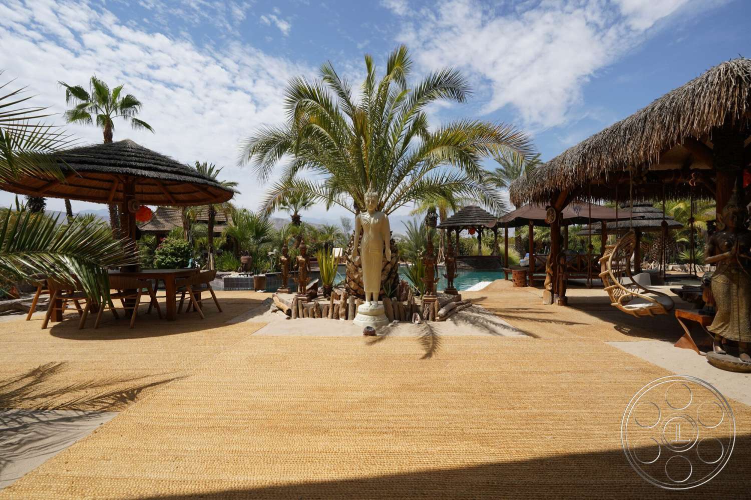 Rancho Mirage 4 - exterior, wooden dining table, woven pendant lights, thatched roof gazebo, palm tree landscape, sunny outdoor environment, tropical garden setting, stone water feature, decorative wooden sculptures, woven grass patio flooring, swinging basket chair, straw beach lounge area, pool adjacent patio, naturalistic stone accents