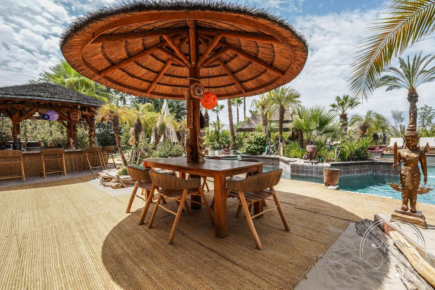 Rancho Mirage 4 - exterior, wooden dining table, outdoor dining area, in-ground swimming pool, sunny outdoor setting, shaded patio space, palm trees landscaping, stone pool border, tropical garden environment, woven rattan dining chairs, decorative garden sculptures, straw palapa gazebo, natural fiber patio flooring