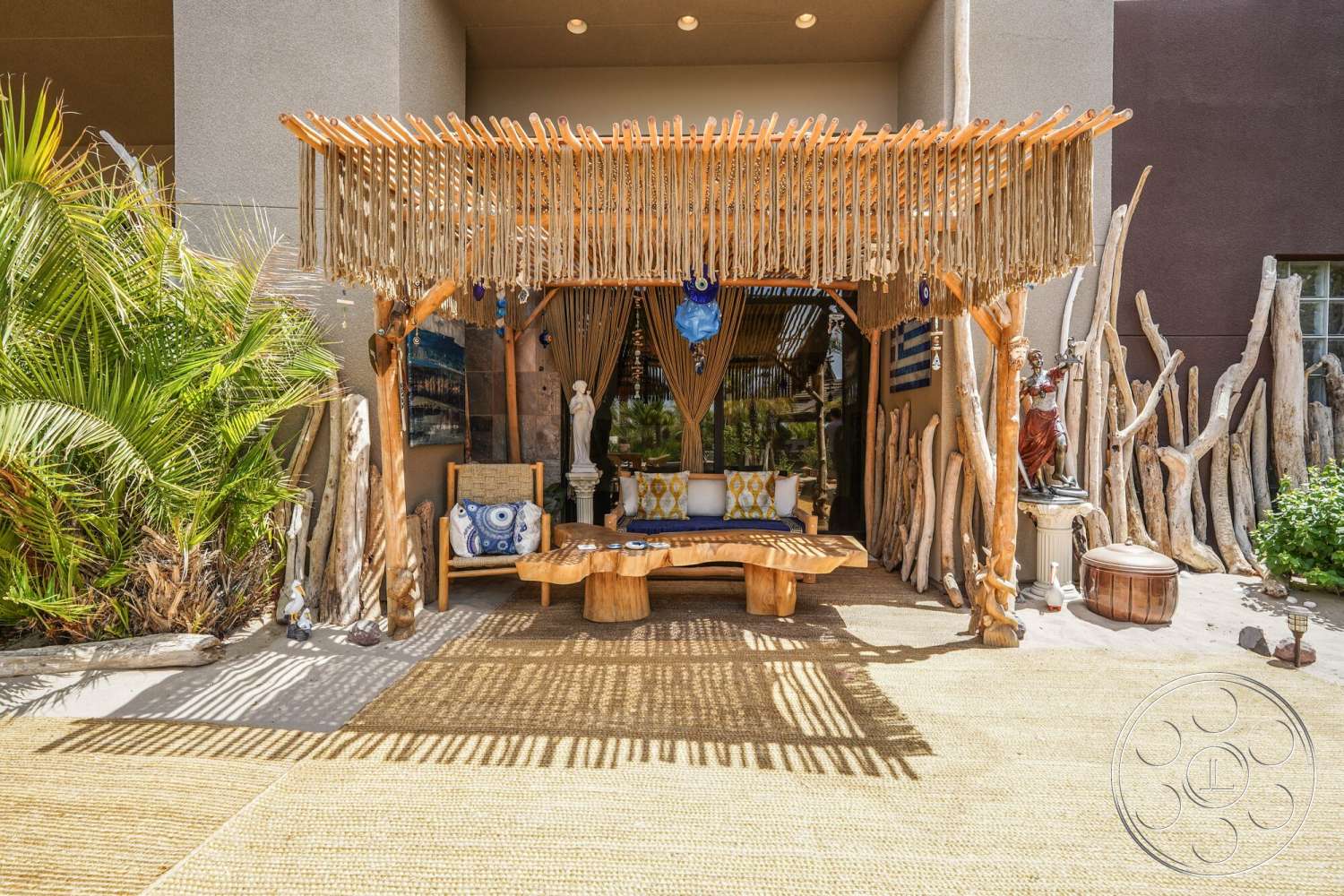 Rancho Mirage 4 - natural wood dining table, potted tropical plants, tropical garden setting, cushioned seating area, patio lounging area, interior transitional space, exposed wood support beams, woven rope detail, decorative stone statues, wooden porch roof structure, woven jute patio flooring, earth-toned exterior walls, bohemian outdoor decor, rustic driftwood accents