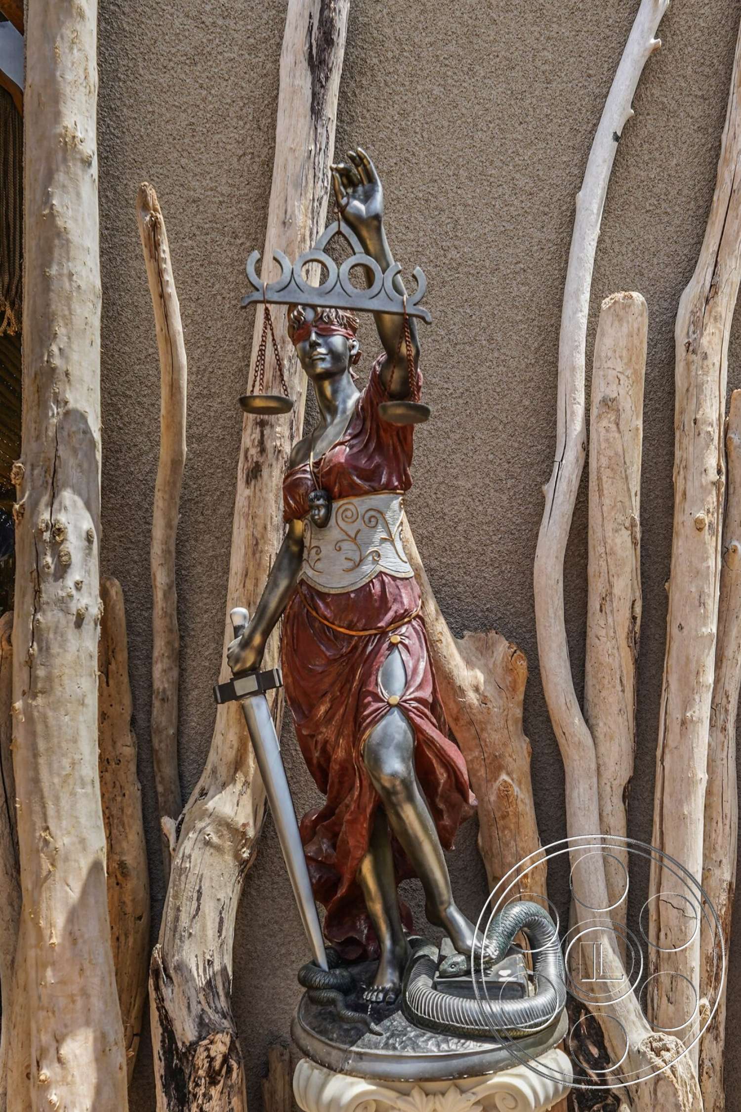 Rancho Mirage 4 - textured stucco wall, weathered wood accents, bronze lady justice statue, smooth stone pedestal base, exterior garden feature, justice scales, sword detail in statue, natural driftwood surrounding