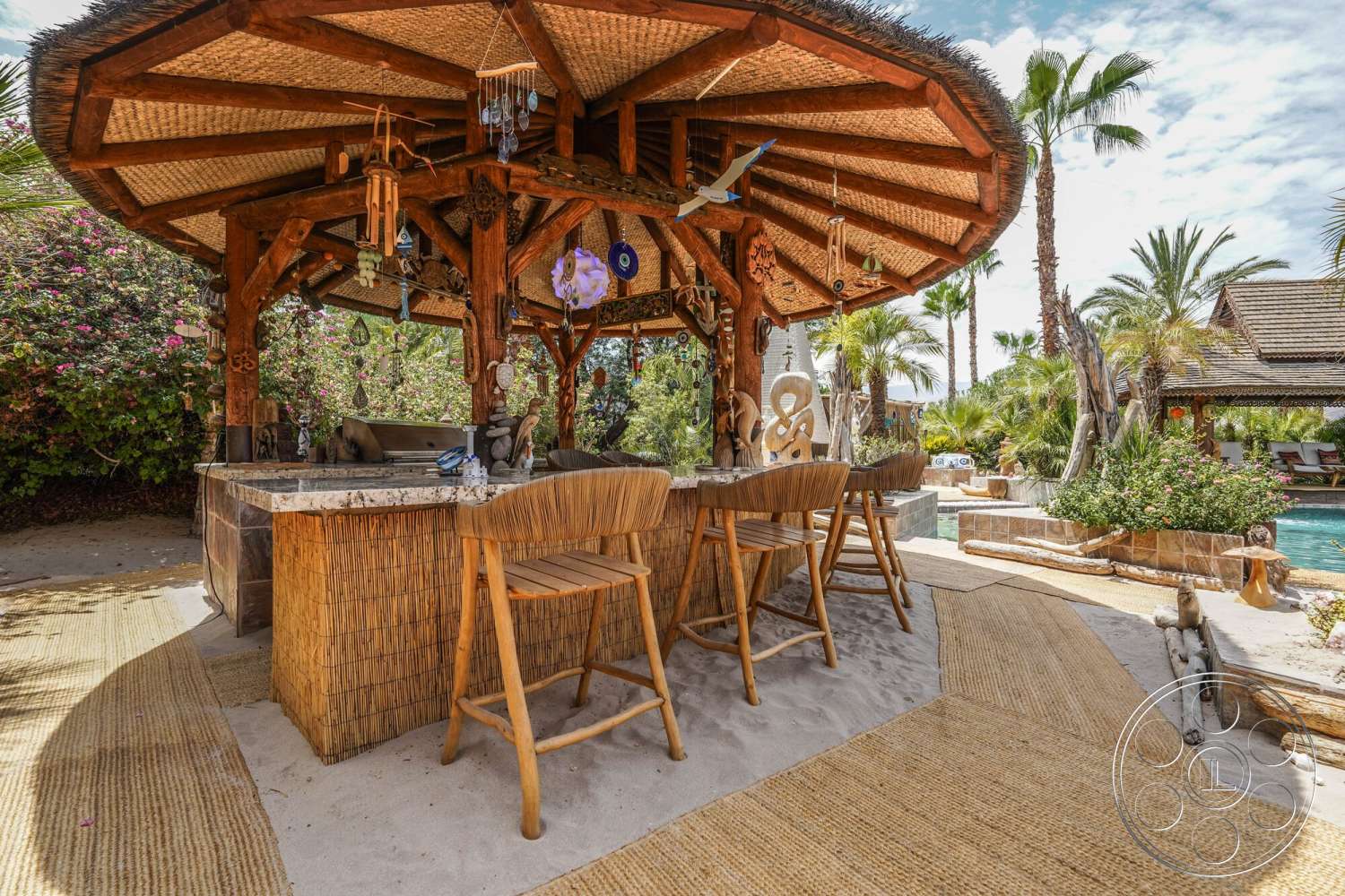 Rancho Mirage 4 - exterior, natural stone accents, woven area rug, tropical landscaping, sand patio flooring, thatched roof patio gazebo, wooden structural beams, granite countertop bar, bamboo bar facade, palms trees surrounding, outdoor bar seating, decorative wind chimes, colorful hanging ornaments, stone outdoor grill, lush floral arrangements