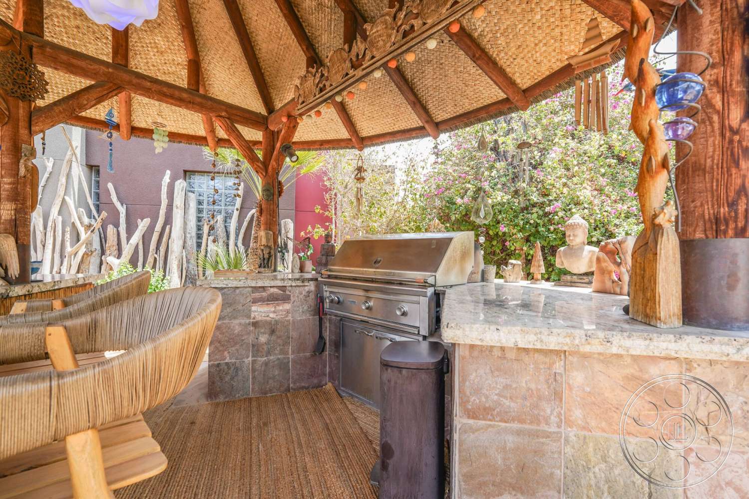 Rancho Mirage 4 - exterior, granite countertop, tropical foliage, outdoor grilling area, landscaped garden backdrop, decorative wooden carvings, thatched roof pavilion, natural wood structure, stone countertop outdoor kitchen, slate stone wall cladding, woven rattan bar stools, textured woven bamboo ceiling, hanging glass ornaments