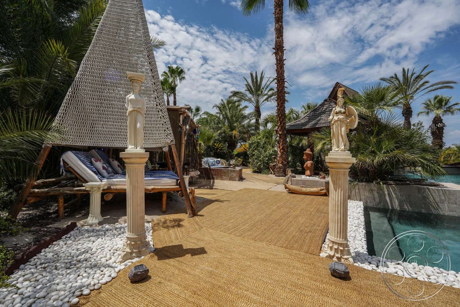 Rancho Mirage 4 - exterior, thatched roof gazebo, infinity pool, tropical palm trees, sunny outdoor space, lush garden setting, decorative stone statues, woven outdoor canopies, beach-style seating area, natural jute patio flooring, white stone pathway border, textured concrete pool edge