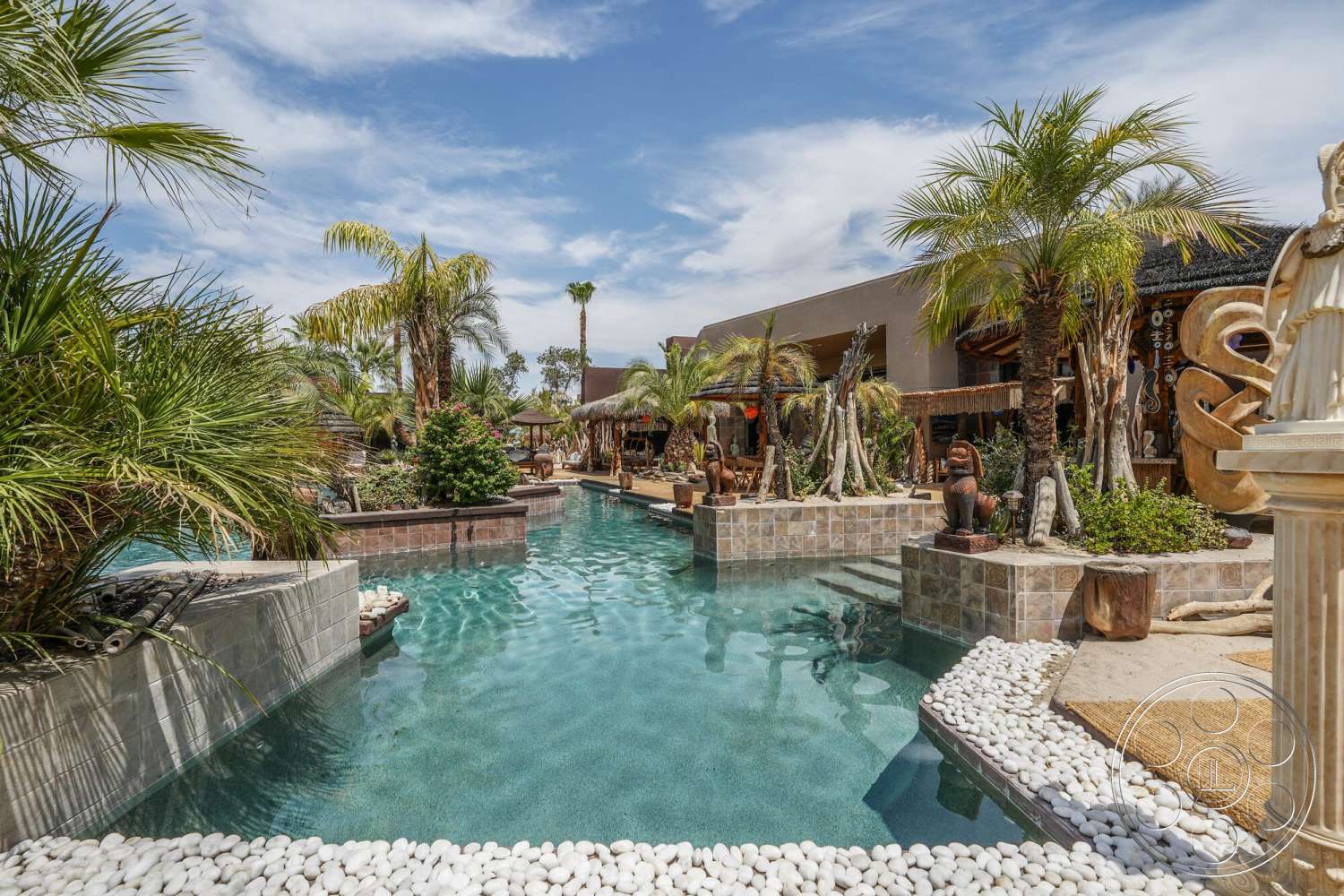 Rancho Mirage 4 - exterior, lush greenery, palm trees, tropical landscaping, wooden pergola, in-ground swimming pool, rectangular pool shape, stone pool deck, sunny backyard setting, natural stone water feature, decorative garden statues, pebble pool border, thatched roof structures, gravel walking paths, outdoor lounge areas, tropical resort style