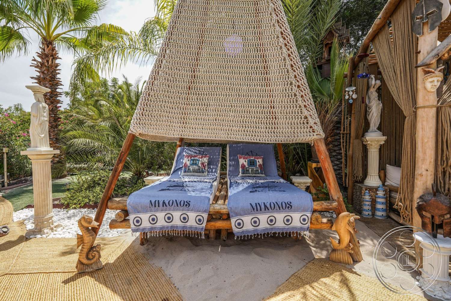 Rancho Mirage 4 - exterior, tropical palm trees, bamboo privacy screen, natural wood lounge chairs, decorative garden sculptures, sand patio flooring, woven canopy structure, textile patterned throw blankets, natural jute area rug, ceramic garden planters