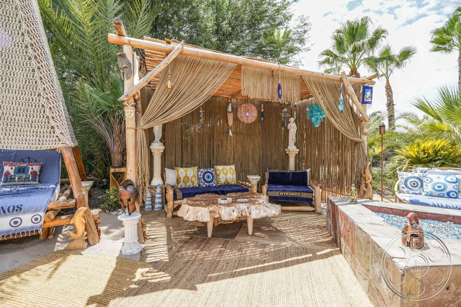 Rancho Mirage 4 - exterior, thatched roof, natural wood furniture, tropical palm trees, woven jute area rug, colorful decorative pillows, textured stone planter, bamboo outdoor structure, woven curtain panels, marble-topped side table, plumeria surrounding, ceramic garden decor, circular stone water feature, handcrafted wooden benches, coastal-themed accents