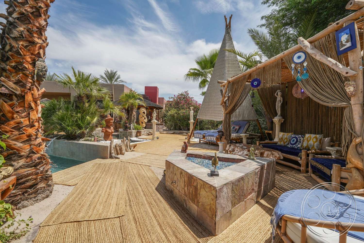 Rancho Mirage 4 - exterior, palm trees, decorative sculptures, tropical landscaping, outdoor living area, concrete pool, concrete pool border, natural stone fire pit, wooden lounge seating, thatched roof structure, woven straw patio flooring