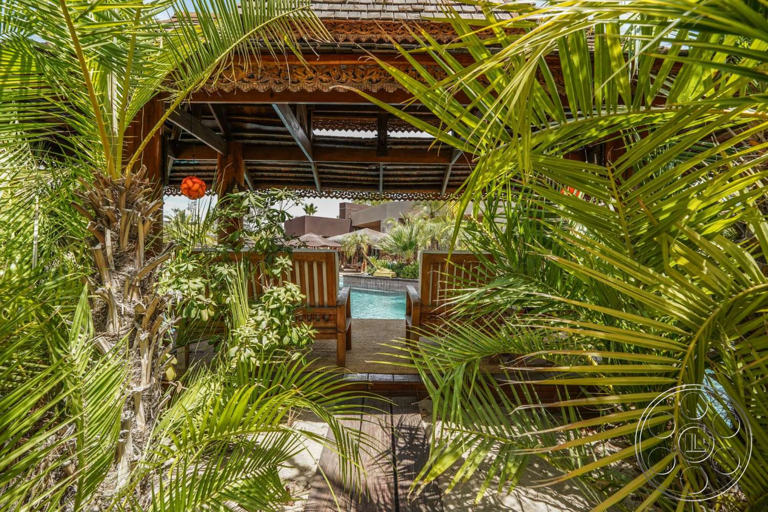 Rancho Mirage 4 - exterior, rectangular swimming pool, thatched roof, stone pool deck, concrete patio floor, palm tree landscaping, wooden seating area, lush tropical foliage, wooden gazebo structure, decorative carved wood details, overhead ceiling beams
