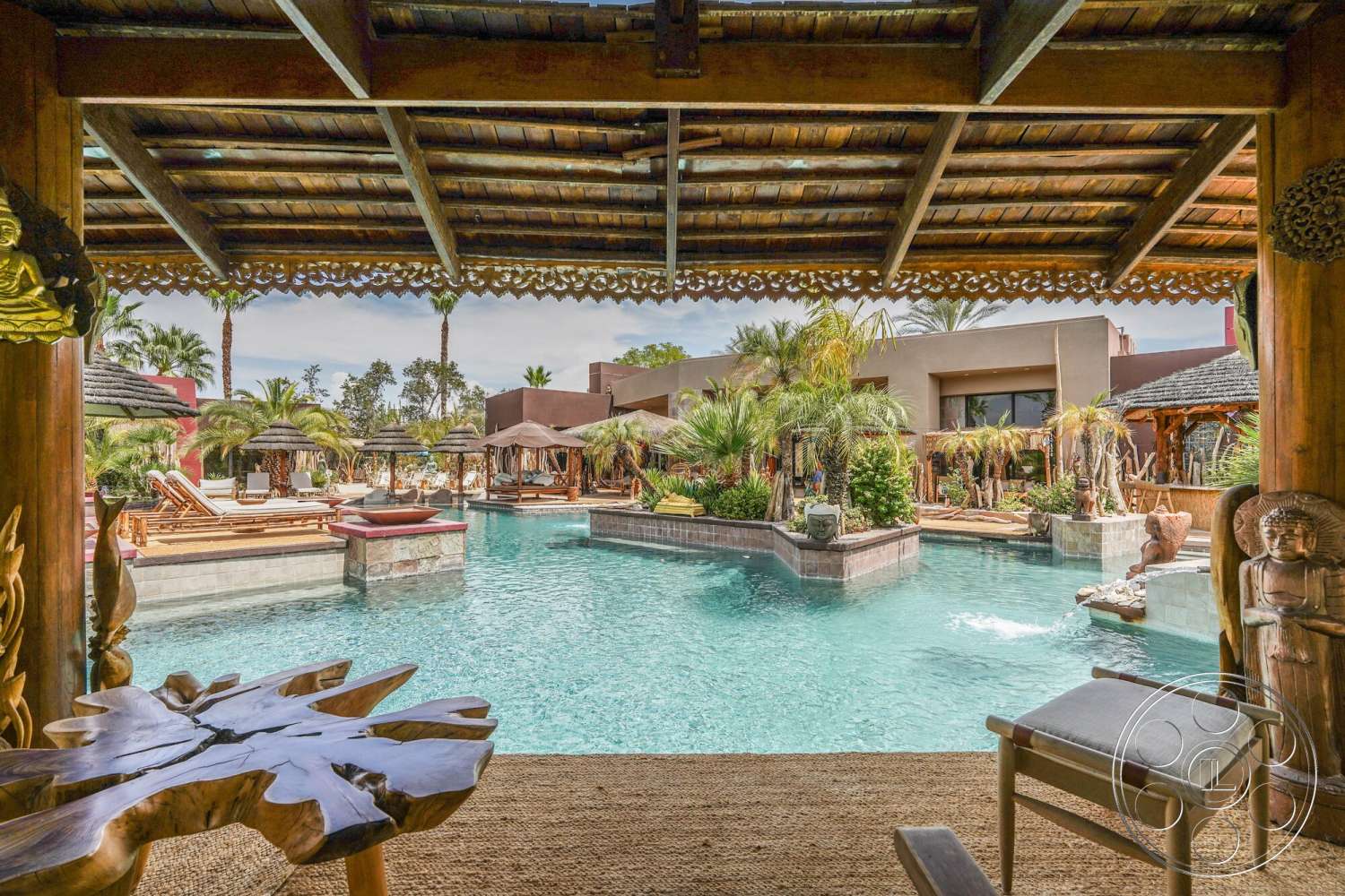Rancho Mirage 4 - exterior, paved pathways, textured plaster walls, infinity pool, palm trees landscaping, natural stone pool deck, open outdoor setting, decorative stone sculptures, wooden poolside loungers, thatched roof pergola, outdoor cabanas, colorful tropical plants, decorative wood railing, modern backyard oasis