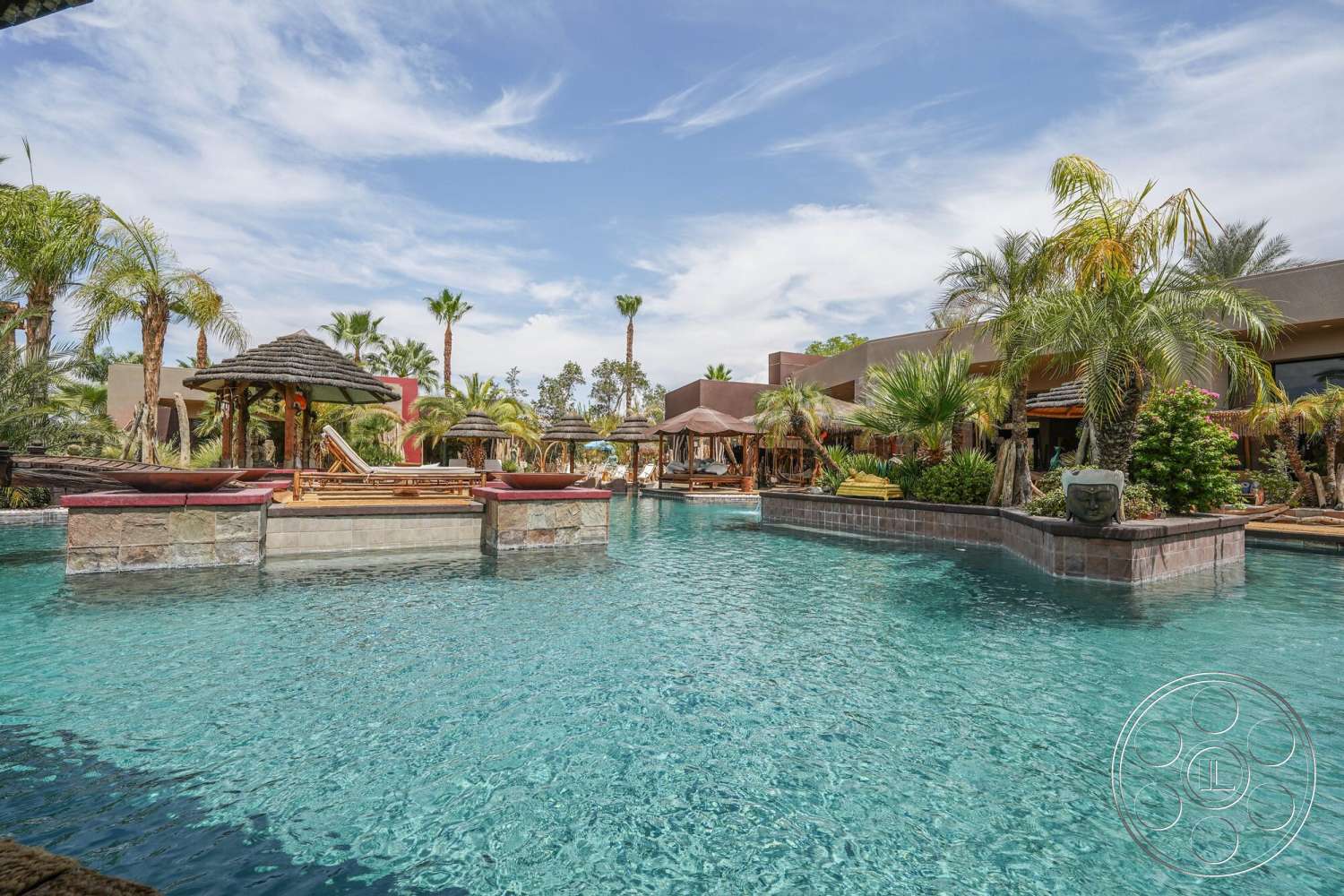 Rancho Mirage 4 - exterior, landscaped garden, palm trees, outdoor seating area, clear blue sky, infinity pool, tropical setting, wooden lounge chairs, stone pool deck, thatch roof cabanas, brick water feature