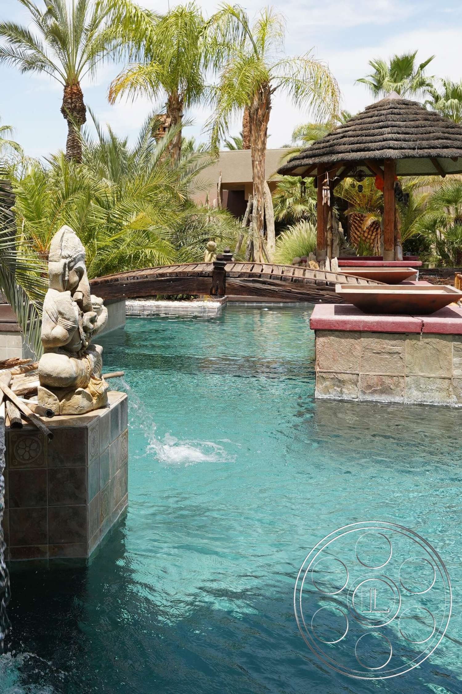 Rancho Mirage 4 - exterior, oval swimming pool, palm tree landscaping, natural stone pool deck, tiled water feature, wooden footbridge, thatched gazebo roof, carved stone statue, lagoon-style pool setting, granite countertop for decorative basin