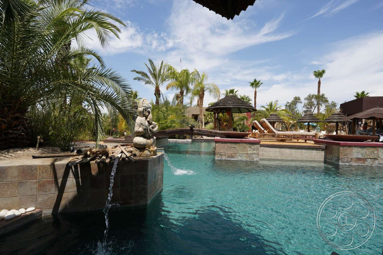 Rancho Mirage 4 - exterior, palm trees, outdoor lounge area, tropical landscaping, sun loungers, stucco walls, rectangular pool, natural stone pool deck, concrete patio border, tiled water feature, decorative stone sculpture, thatched roof cabanas, ornamental stone wall