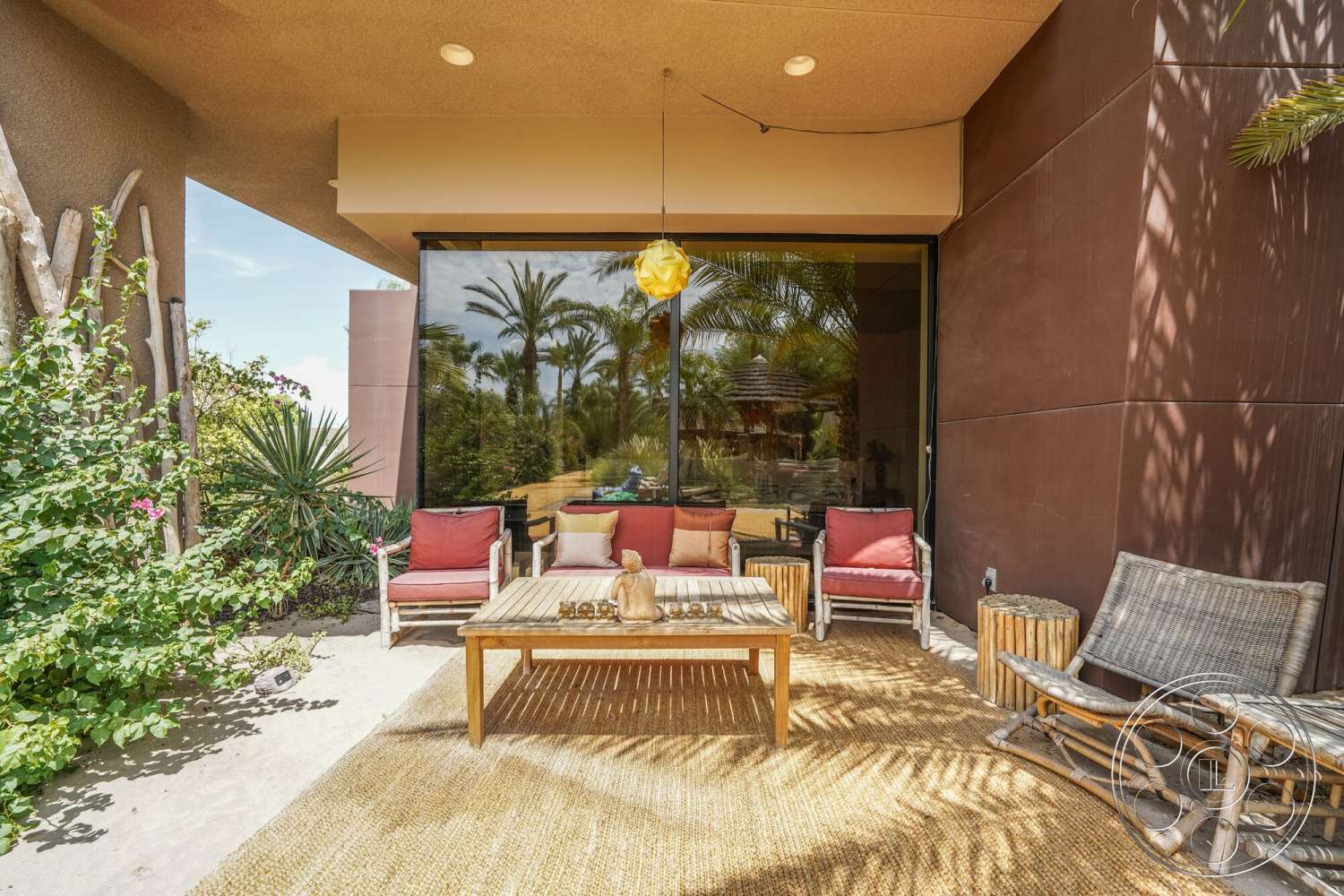 Rancho Mirage 4 - exterior, wooden coffee table, glass sliding doors, modern pendant light fixture, stucco wall finish, decorative stone accents, concrete patio floor, tropical landscaping with palm trees, woven natural fiber area rug, upholstered chairs with red cushions, reclining wooden lounge chair, potted plants and shrubs