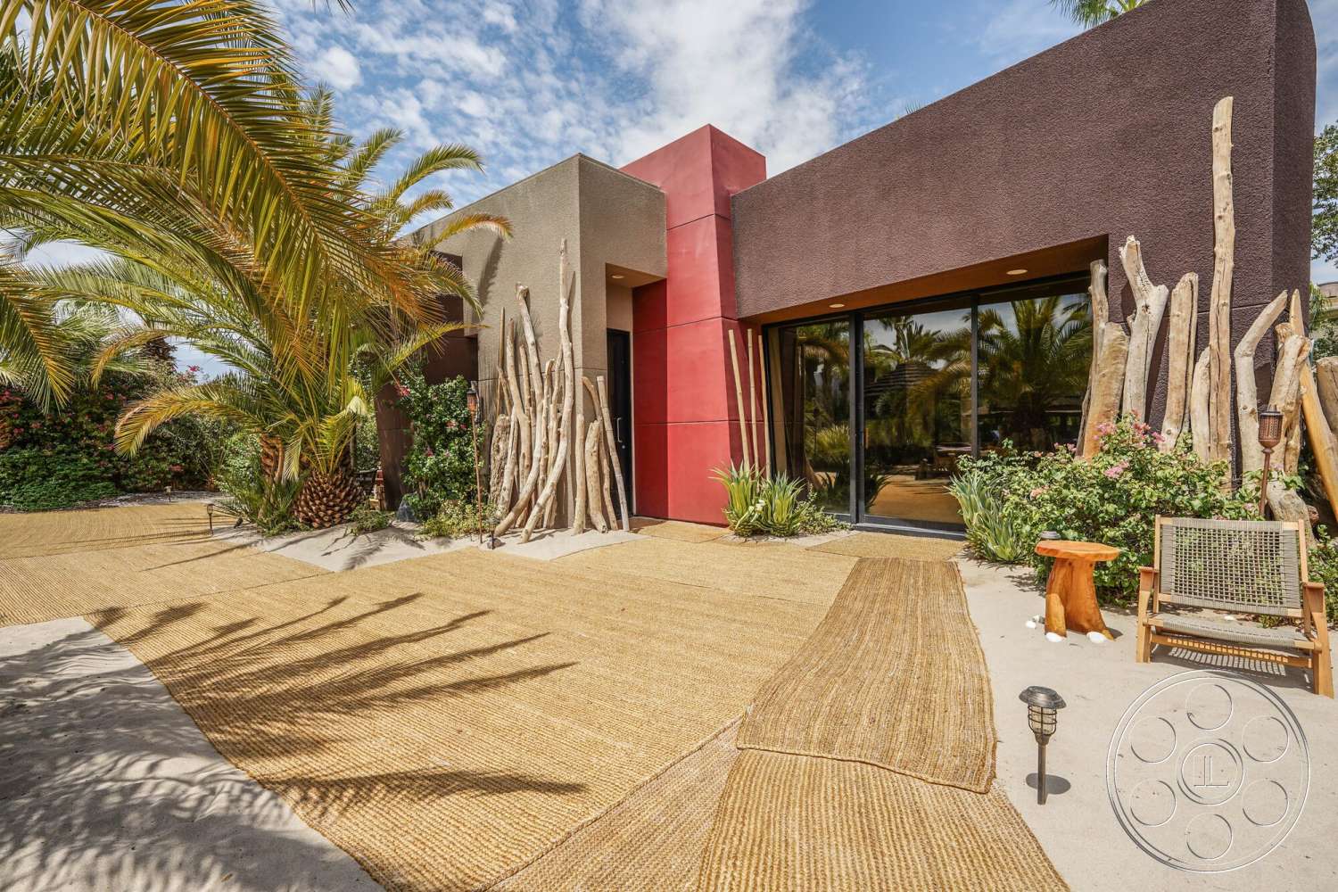 Rancho Mirage 4 - exterior, red accent wall, gravel pathway, modern architectural style, wooden lounge chair, palm tree landscaping, floor-to-ceiling glass doors, tropical garden setting, stucco wall facade, natural wood features, woven outdoor rugs, sandstone patio, bamboo garden features