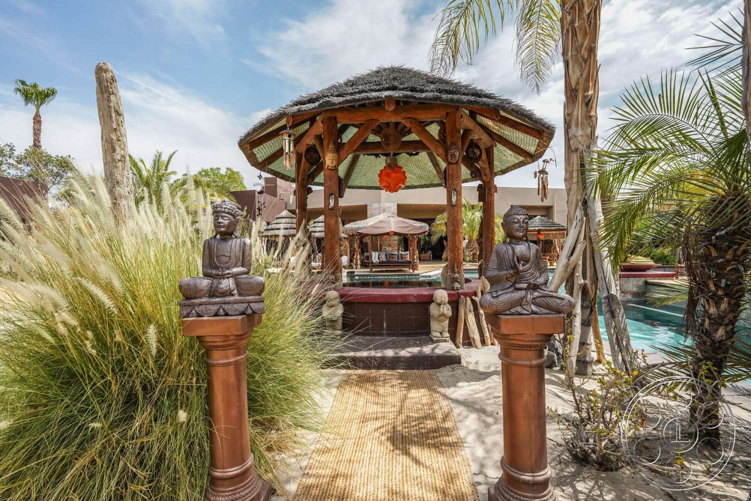 Rancho Mirage 4 - exterior, thatched roof, tropical palm trees, drought-resistant landscaping, desert home setting, decorative stone statues, wooden support columns, bamboo gazebo structure, substantial ornamental grasses, stone pool edges, stone water features, woven outdoor rug, relaxation area with lounge seating, open-air summer space