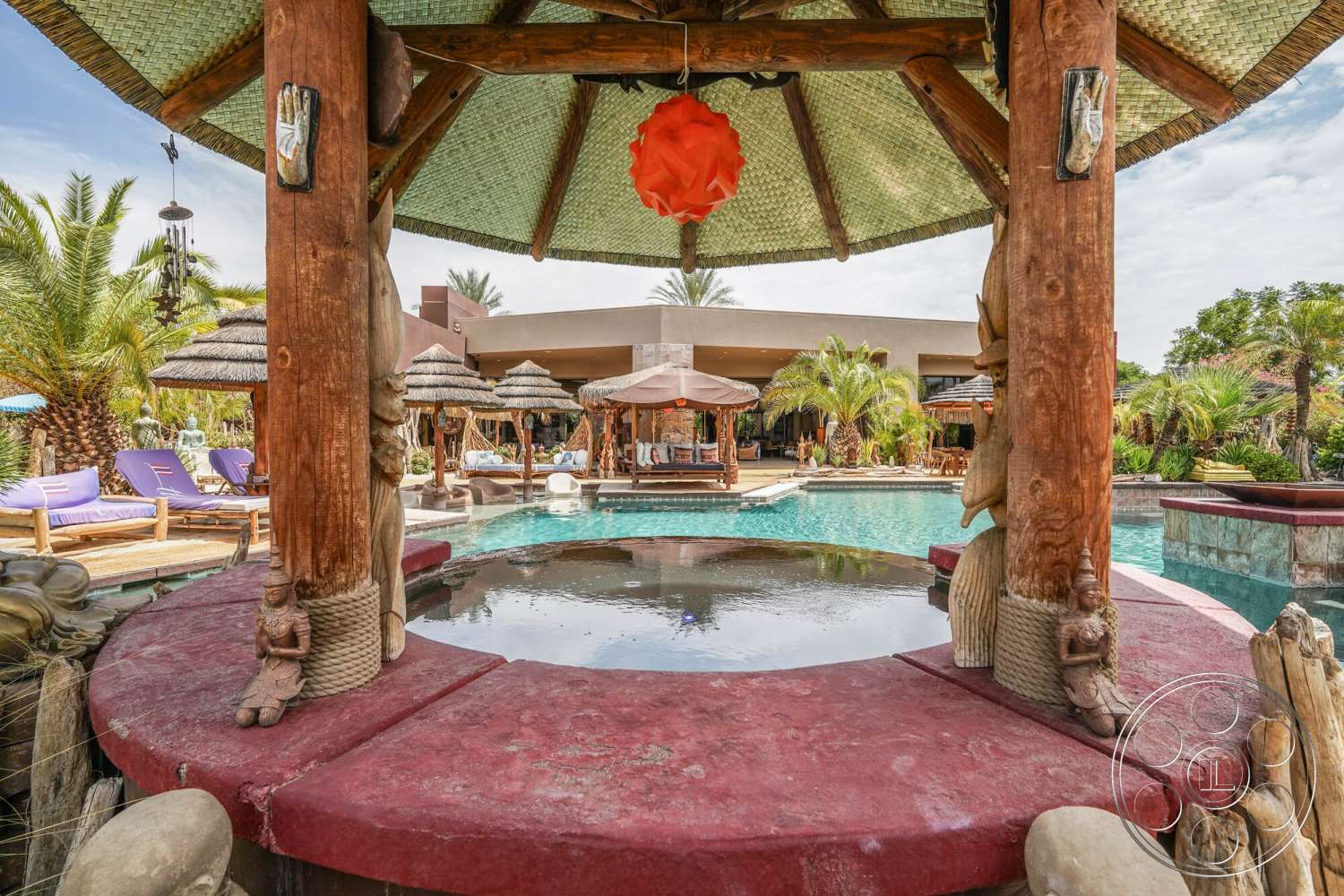 Rancho Mirage 4 - exterior, tropical landscaping, thatched roof gazebo, natural stone pool deck, concrete pool surround, palm tree setting, open-air gazebo, wooden support columns, pool area with water features, red plastered pool bar, outdoor lounge with purple sun loungers, sculptural stone elements, mandala-style decorative elements, desert oasis atmosphere