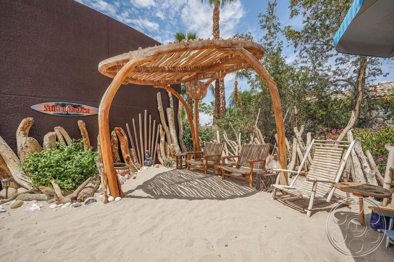 Rancho Mirage 4 - exterior, sunny outdoor setting, sand flooring, landscaped garden area, wooden pergola structure, beach-themed decor, lush green plants, natural wood seating, decorative driftwood accents, tropical environment