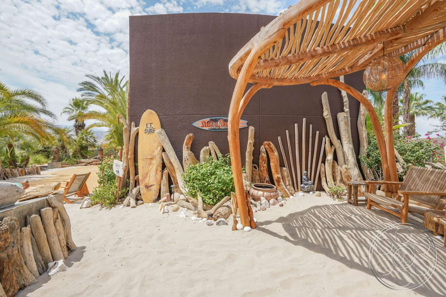 Rancho Mirage 4 - exterior, lush greenery, outdoor seating area, desert setting, palm tree landscaping, stone water feature, weathered wood accents, thatched roof structure, natural wood seating, textured brown wall, driftwood art installation, sand beach area, surfer-style wooden surfboard