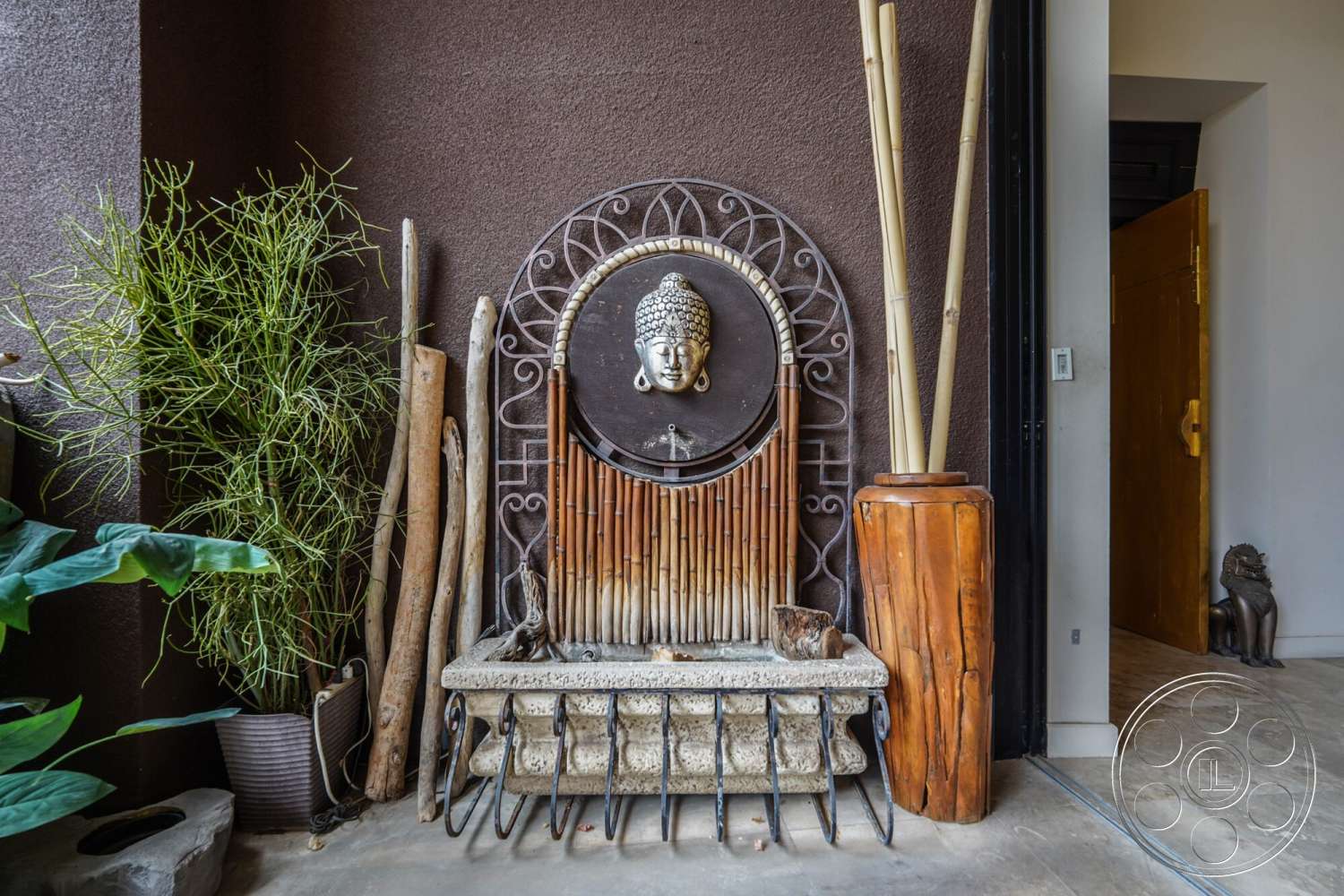 Rancho Mirage 4 - exterior wall treatment with brown textured plaster, stone water feature with bamboo accents, wooden planter with tropical foliage, decorative bamboo poles, natural wood sculptures, interior entrance with minimalist design, concrete flooring with a polished finish