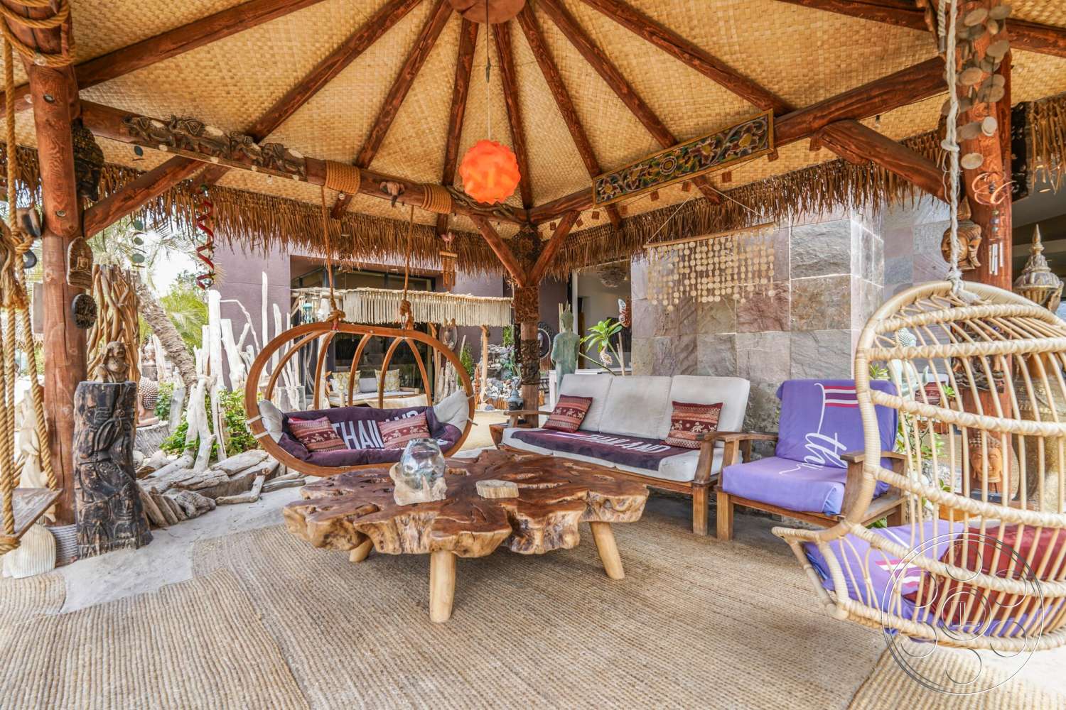 Rancho Mirage 4 - textured area rug, cozy seating arrangement, exposed wooden beams, natural wood furniture, interior outdoor living space, modern bohemian style, tropical outdoor setting, woven thatched roof, organic shaped coffee table, hand-woven seating cushions, hanging rattan chair, decorative stone wall backdrop, open air lounge area