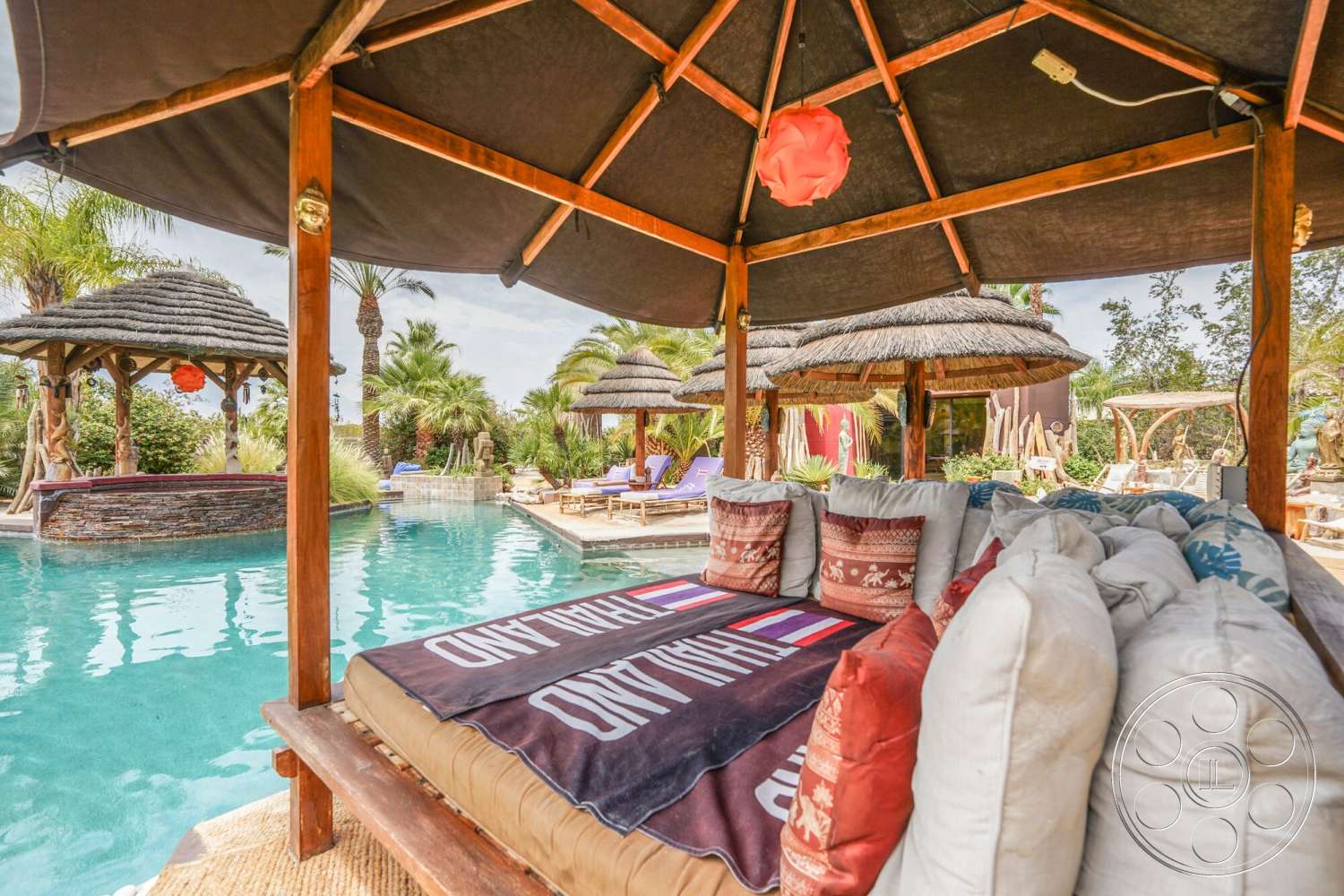 Rancho Mirage 4 - exterior, palm trees, tropical landscaping, thatched roof gazebo, wooden structure, infinity pool, decorative throw pillows, textured fabric cushions, poolside lounge area, stone pool deck, woven outdoor rugs