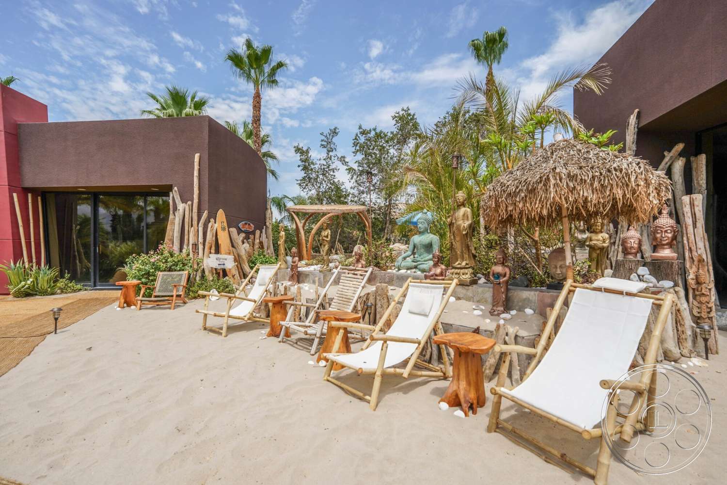 Rancho Mirage 4 - exterior, palm trees, red accent wall, natural wood elements, outdoor lounge area, thatched roof gazebo, stucco walls, wooden support beams, decorative stone sculptures, wooden beach chairs, bamboo side tables, sand patio surface, tropical garden landscaping