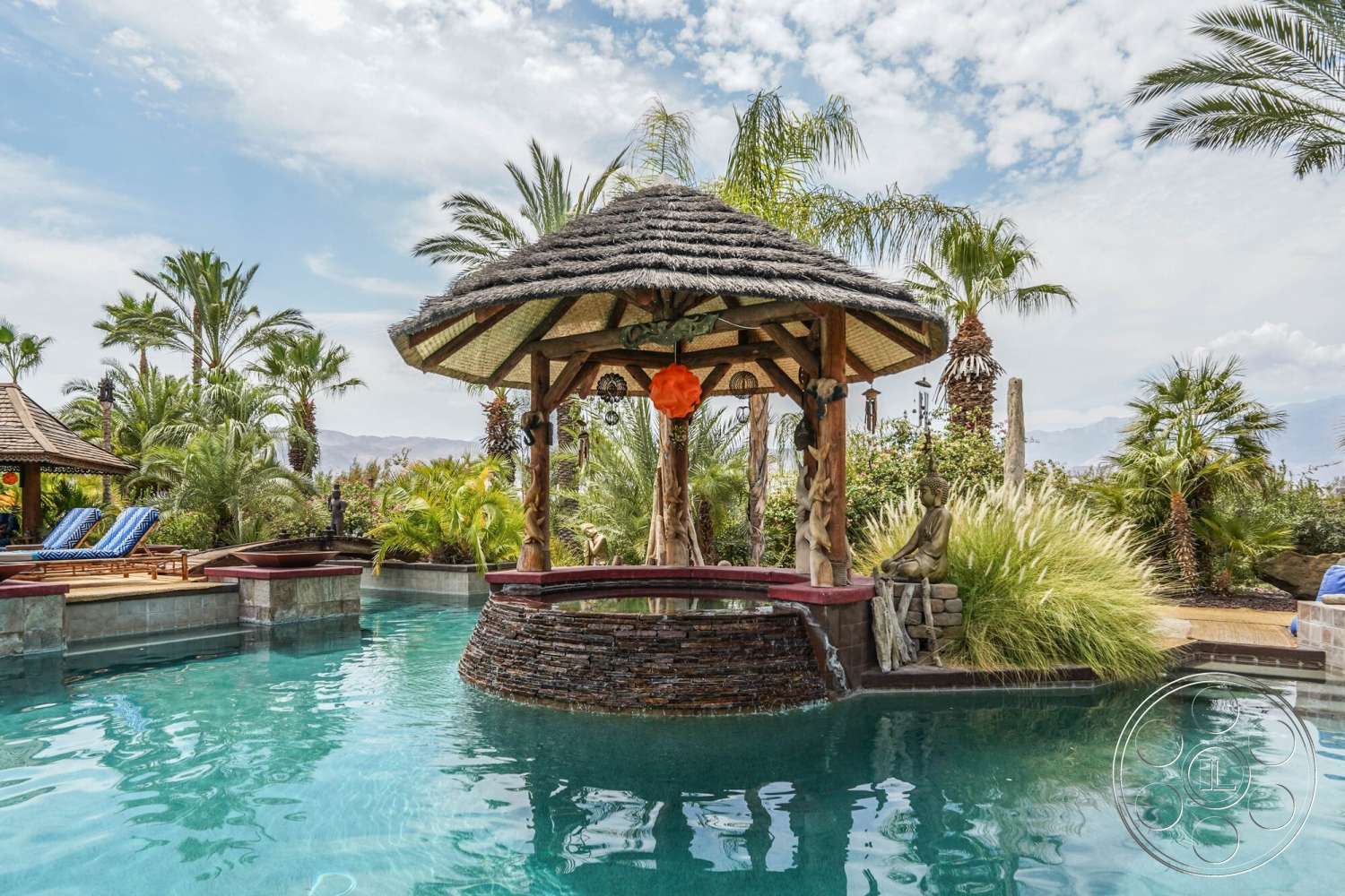 Rancho Mirage 4 - exterior, landscaped garden, natural stone accents, lush greenery, water feature, thatched roof gazebo, infinity pool, tropical palm trees, outdoor lounge chairs, decorative stone wall, stone pool deck, wooden support beams, decorative Buddha statue