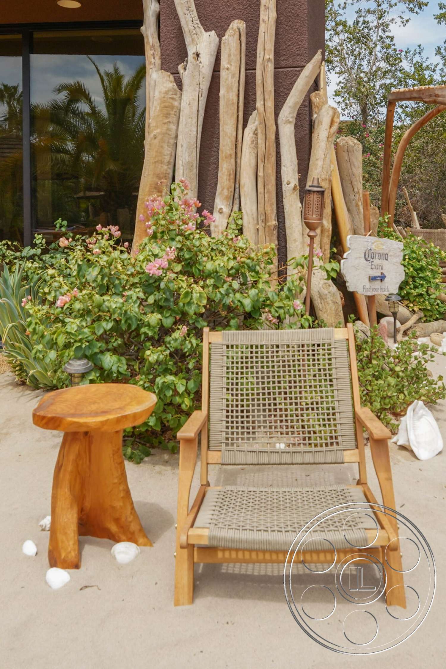 Rancho Mirage 4 - exterior, outdoor lantern, rustic garden setting, natural wood side table, wooden chair with woven fabric seat, weathered wooden tree trunks, green shrubbery with pink flowers, beach sand flooring, decorative seashells