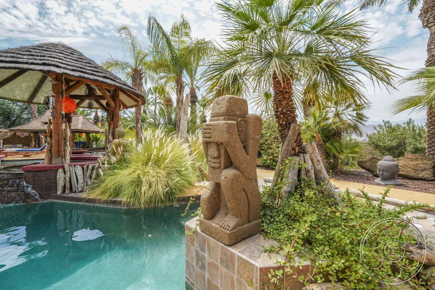 Rancho Mirage 4 - exterior, gravel pathway, thatched roof gazebo, rectangular pool, lush green foliage, decorative stone border, natural stone pool deck, wood frame structure, tropical landscaping with palm trees, textured stone statue, garden area with ornamental grasses