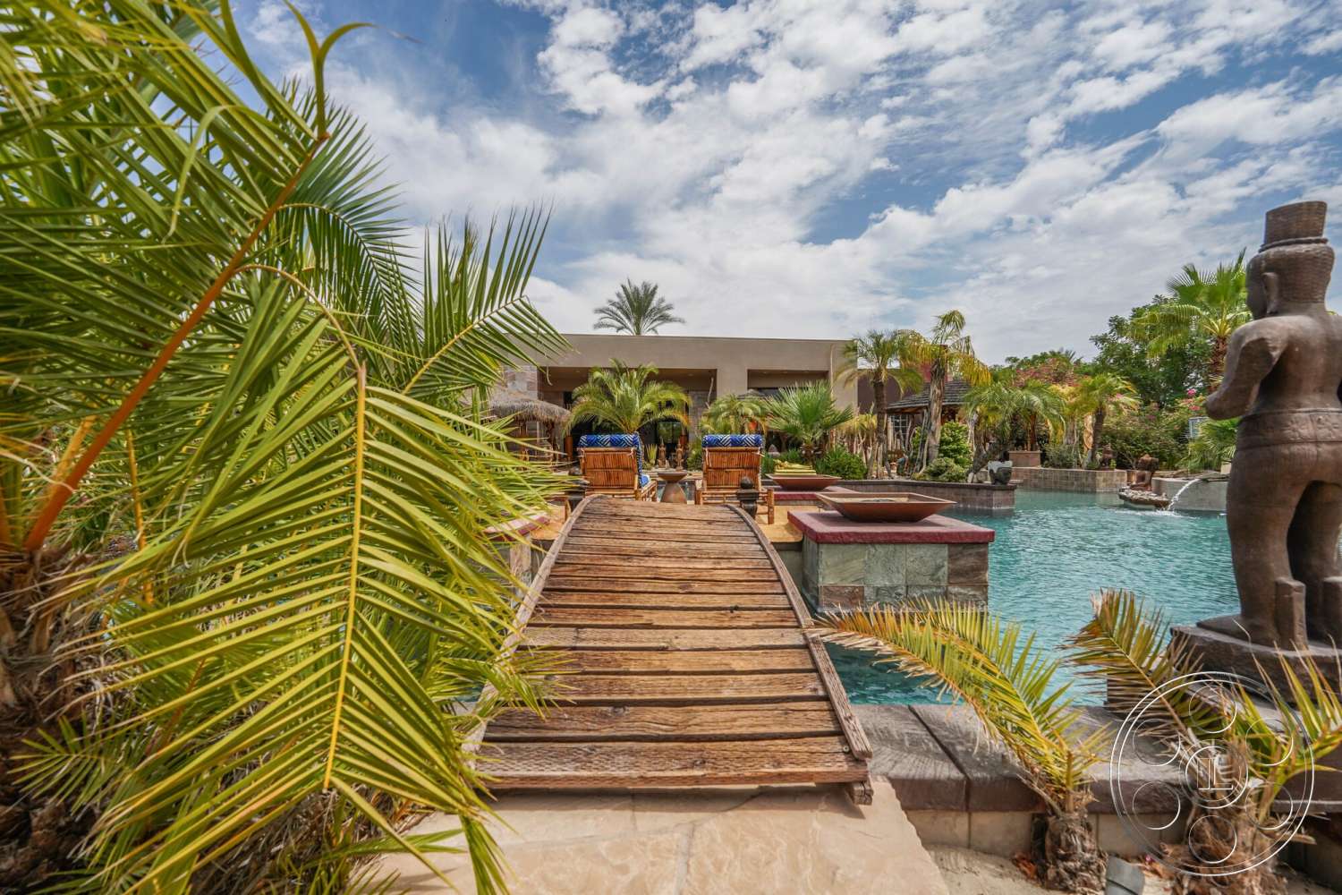 Rancho Mirage 4 - palm trees, potted plants, rectangular swimming pool, tropical landscaping, natural stone steps, sunny outdoor setting, stucco exterior walls, outdoor patio area, stone pool border, decorative stone sculpture, wooden bridge walkway, textured stone features