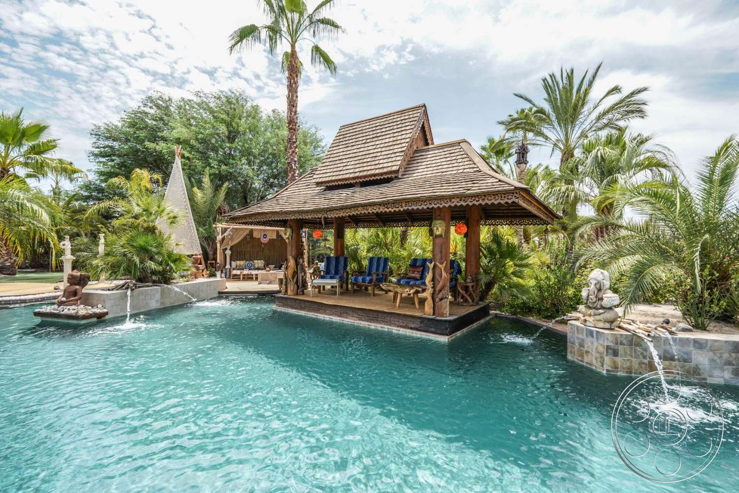 Rancho Mirage 4 - exterior, lush greenery, outdoor seating area, decorative lanterns, tropical landscaping, natural stone wall, concrete pool deck, rectangular pool, wooden patio, stone water feature, palm trees surrounding, thatched roof pavilion, wooden structural supports, sculptural garden decor