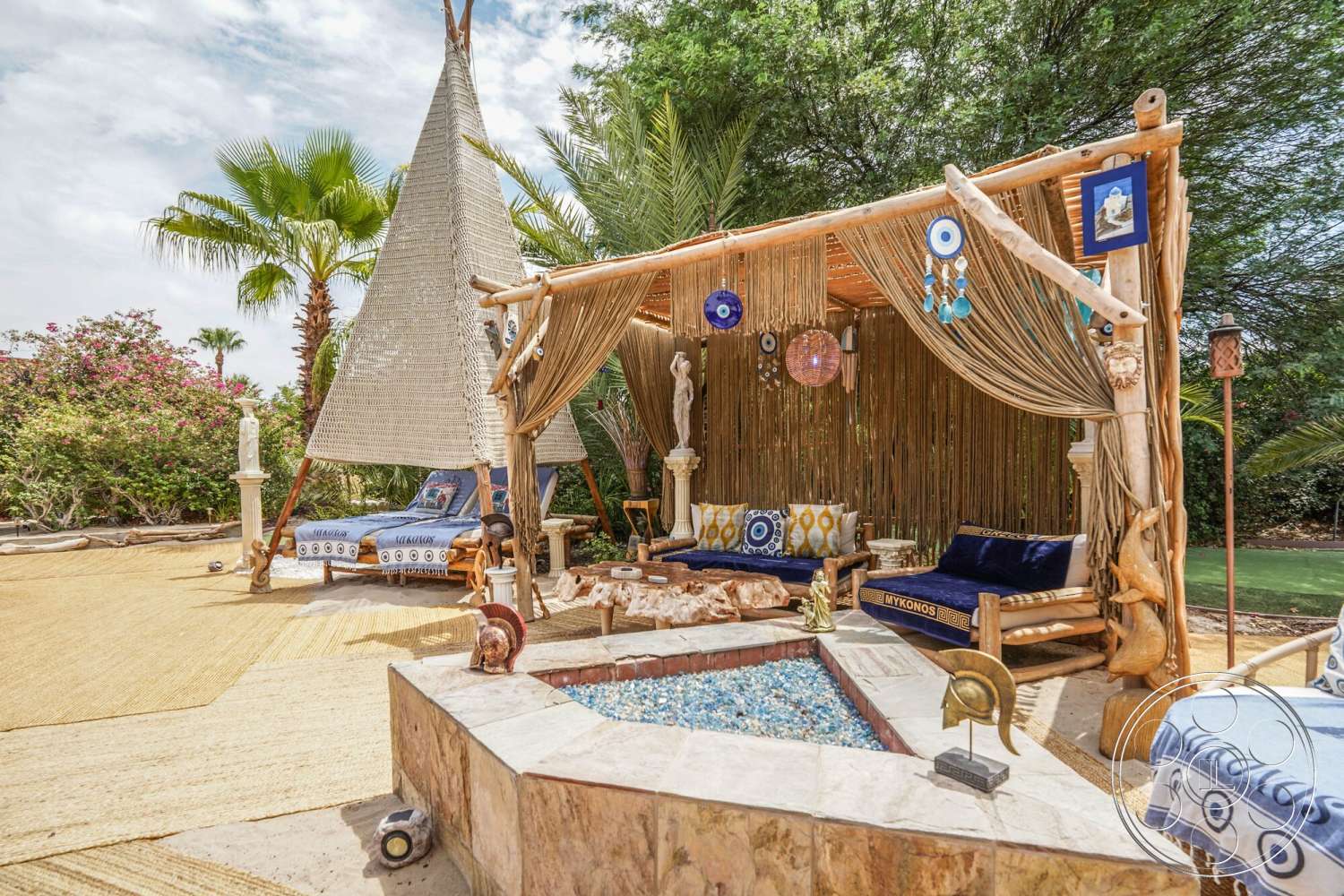 Rancho Mirage 4 - exterior, natural stone accents, decorative wall art, textured outdoor rug, stone fire pit, landscaped garden with tropical plants, relaxed outdoor seating area, wooden lounge seating, woven canopy structure, decorative blue and white cushions, woven hanging curtains