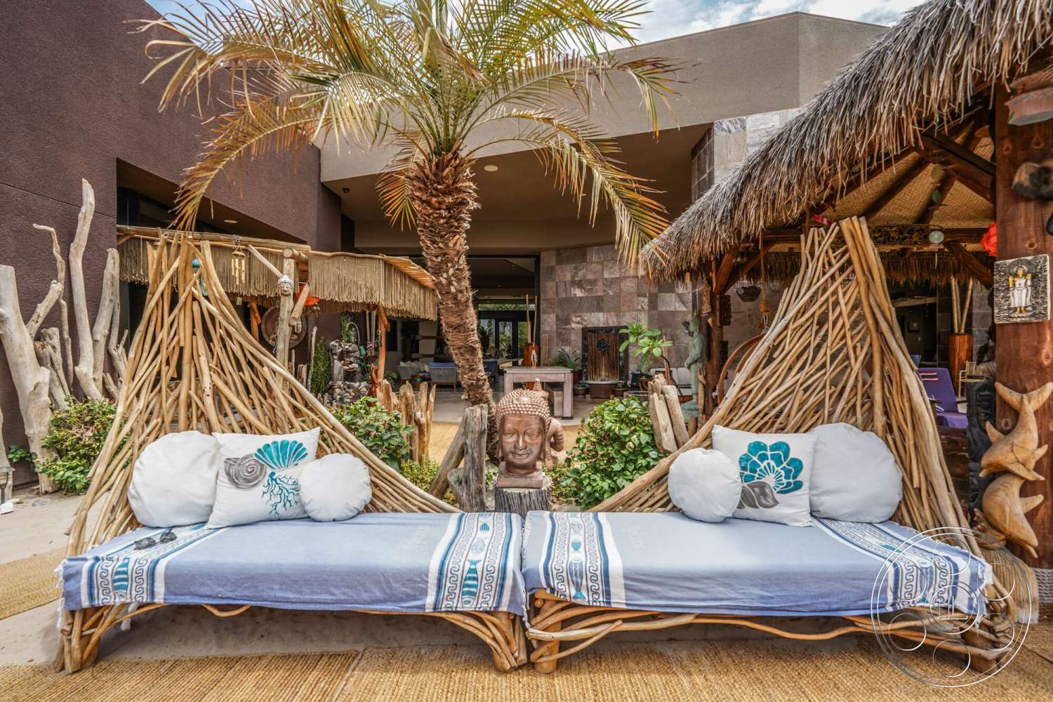 Rancho Mirage 4 - exterior, outdoor living space, textured stone flooring, open patio area, thatched roof structures, woven bamboo lounge seating, textile cushions with patterns, palm trees in the setting, natural wood decorative accents, tropical landscaping with greenery, earth-toned building walls, rustic art sculptures