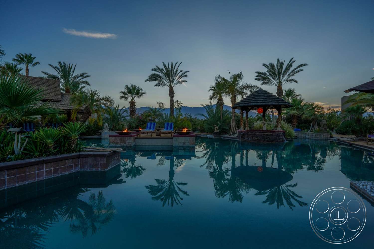 Rancho Mirage 4 - exterior, palm trees, surrounding greenery, mountain view, tropical landscaping, infinity pool, outdoor lounge chairs, reflective water surface, decorative stone walls, twilight setting, stone pool deck, concrete pool border, stone gazebo, outdoor fire features
