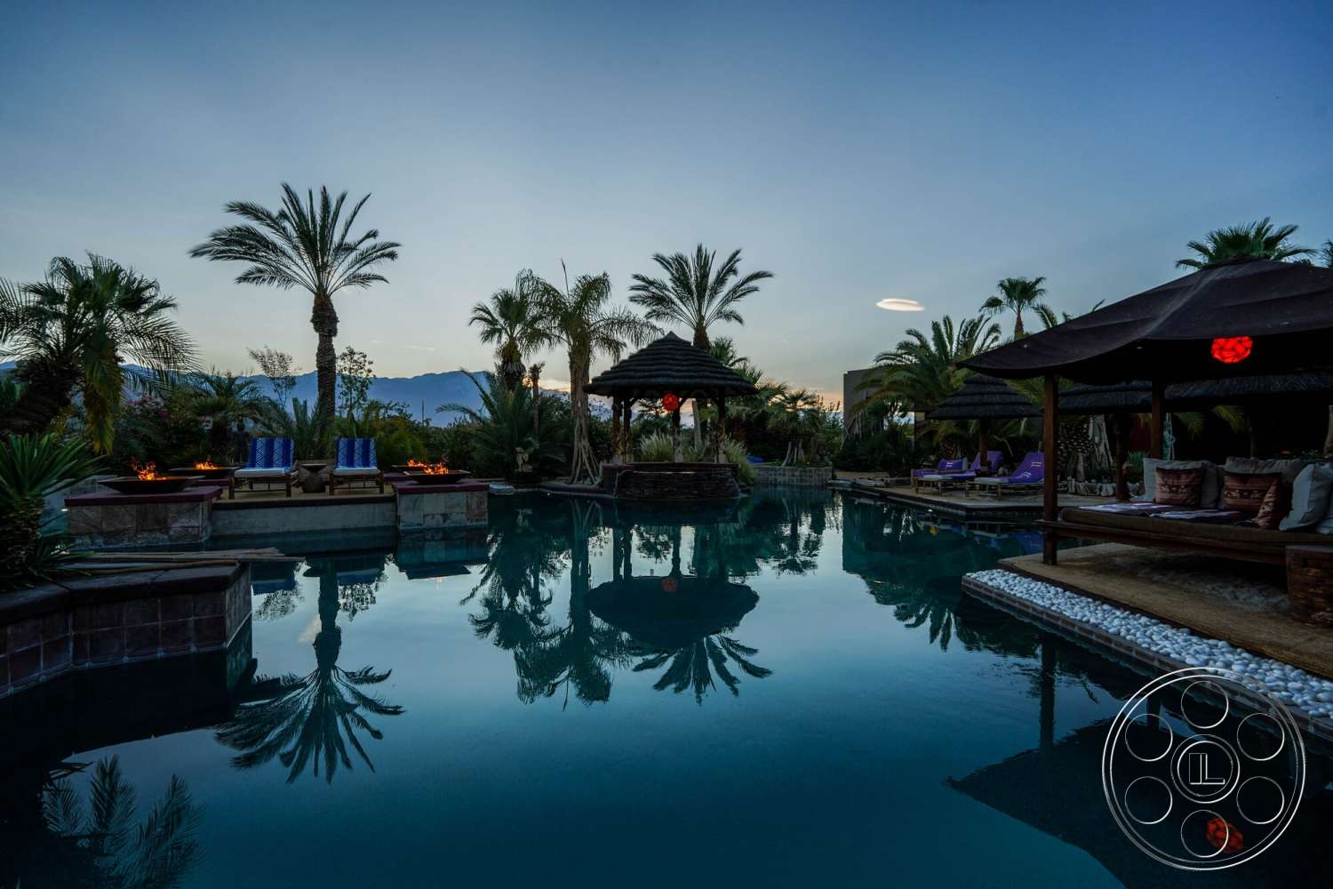 Rancho Mirage 4 - exterior, outdoor lounge area, thatched roof, infinity pool, brick pool deck, palm tree landscaping, mountain view backdrop, natural stone pool border, tropical garden setting, wooden gazebo structure, decorative fire pits, staggered stone wall, water reflection, twilight lighting