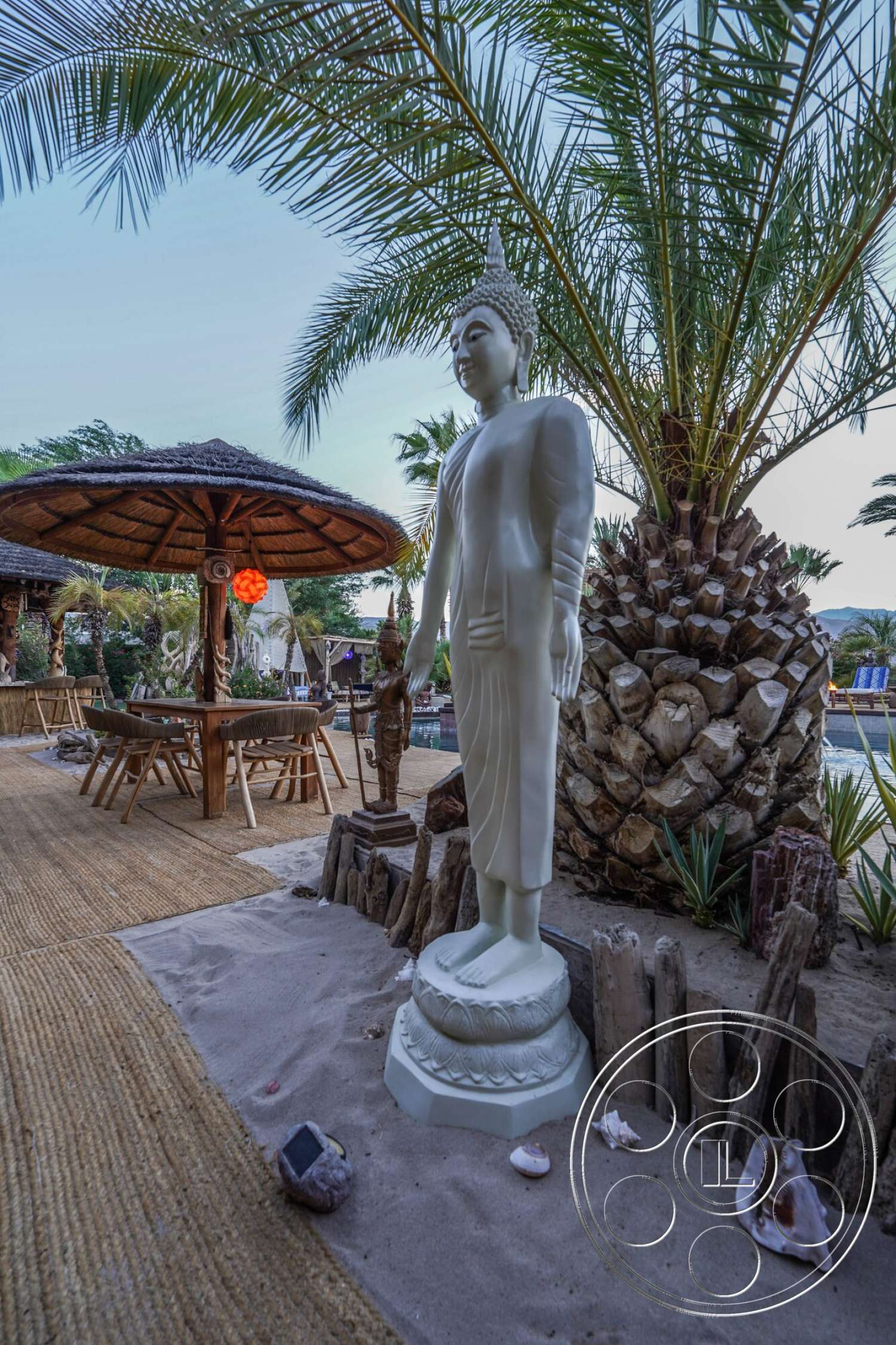 Rancho Mirage 4 - exterior, outdoor seating area, palm tree landscaping, wooden outdoor dining table, beach setting, white marble Buddha statue, thatched roof patio, woven outdoor flooring, sand garden area, native plant decoration