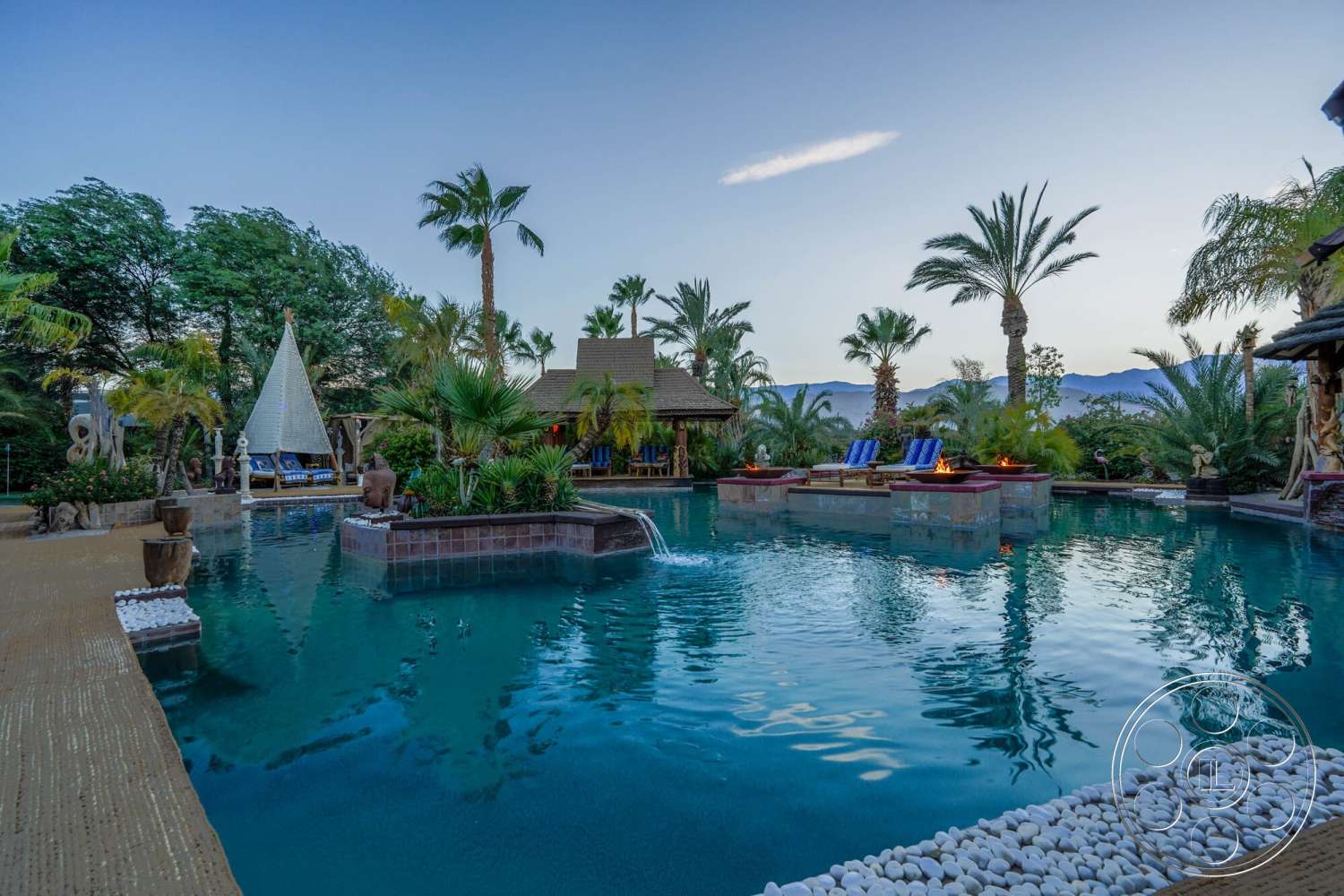 Rancho Mirage 4 - exterior, natural stone accents, palm trees, wooden deck, mountain view, tropical landscaping, outdoor lounging area, concrete pool deck, rectangular pool, fire pit area, thatched roof structure, sculptural garden features, pool with stone coping, white gravel border