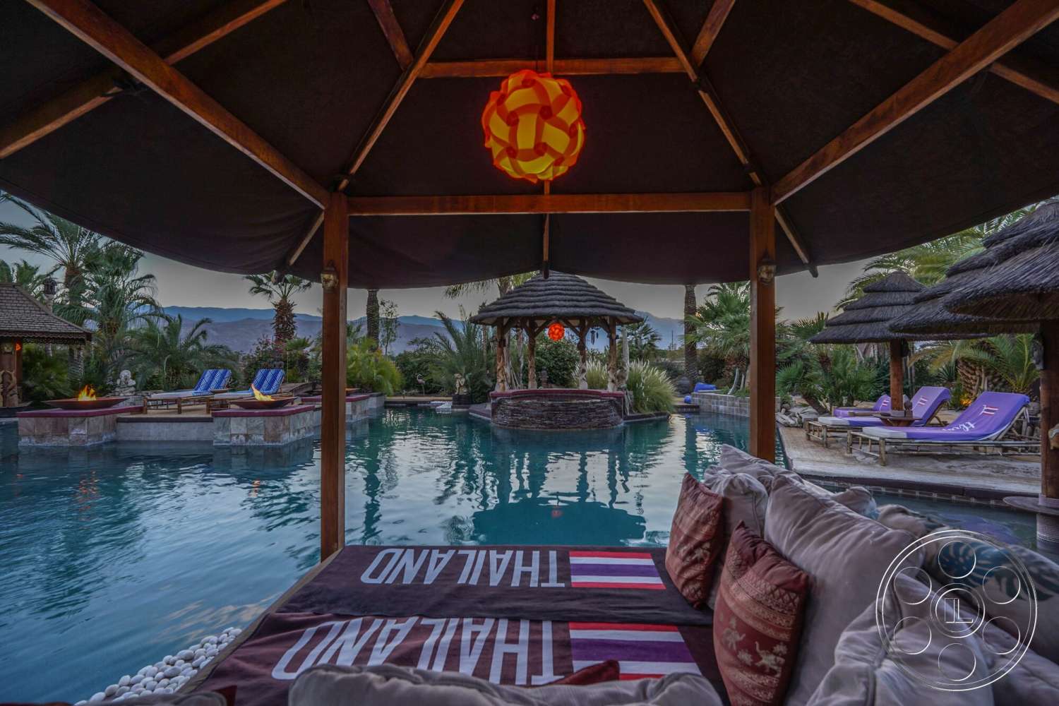 Rancho Mirage 4 - exterior, palm trees, tropical landscaping, thatched roof, wooden lounge chairs, stone pool border, rectangle infinity pool, smooth water surface, wood pavilion structure, outdoor cushions, decorative lantern light, textured outdoor throw blanket