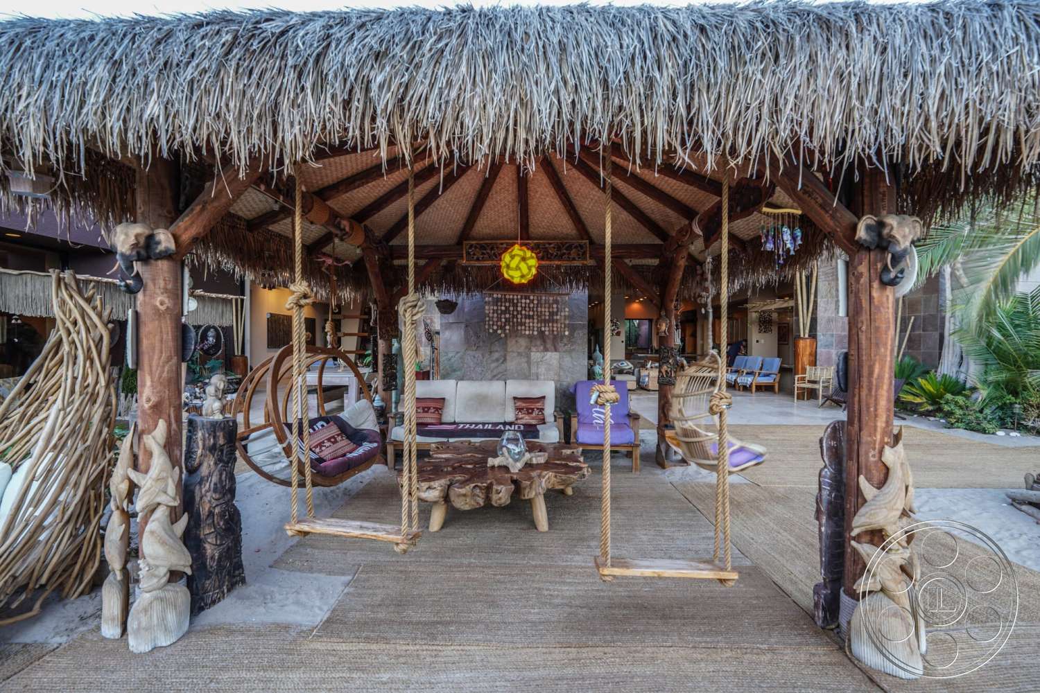 Rancho Mirage 4 - natural stone flooring, outdoor lounge area, open-air design, thatched roof gazebo, interior patio, exposed wooden ceiling beams, wooden support beams, rustic wooden coffee table, tropical garden setting, carved wooden accents, woven seating area, woven swing chairs
