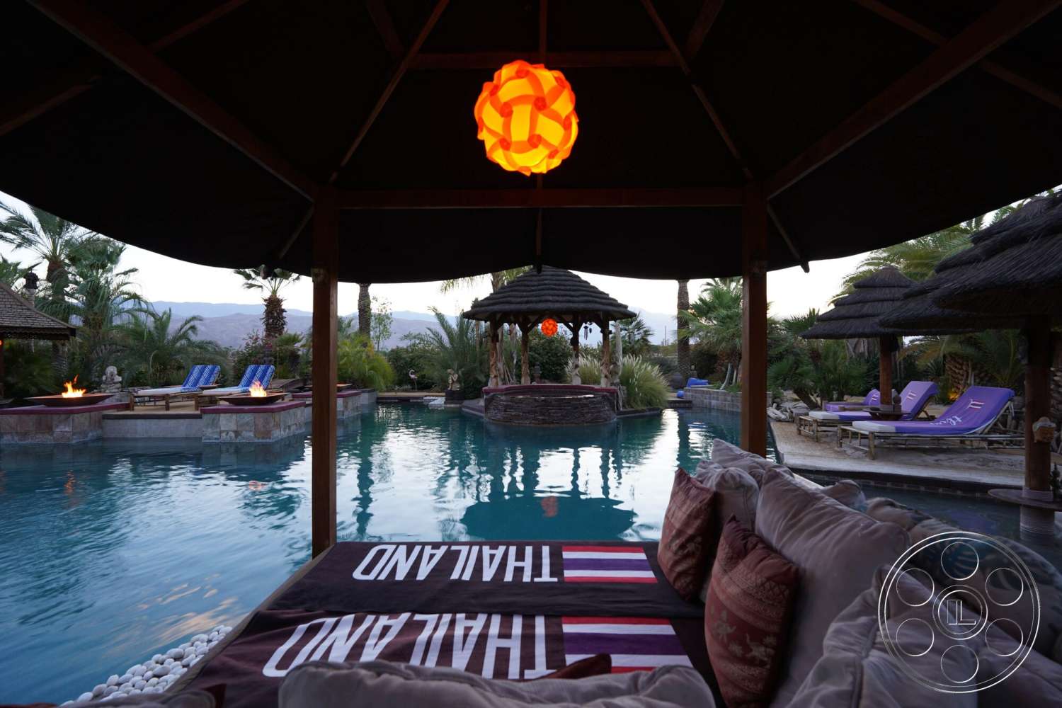 Rancho Mirage 4 - landscaped garden, thatched roof cabana, tropical palm trees, rectangular pool, textured stone walls, wooden lounge chairs, stone pool deck, exterior pool area, modern outdoor living space, fire pit seating area, woven outdoor cushions, outdoor pendant lighting, serene desert backdrop