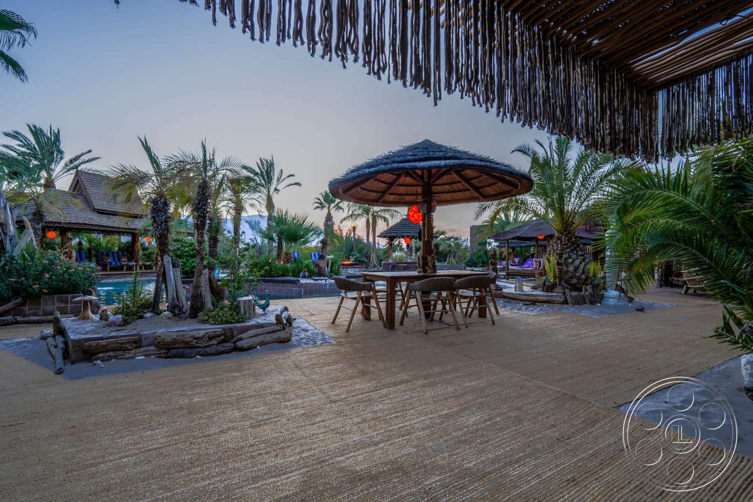 Rancho Mirage 4 - exterior, wooden dining table, outdoor living space, thatched gazebo, palm trees landscaping, exposed wood ceiling, natural stone pool border, tropical garden setting, pool area with water feature, woven seating chairs, sandy patio flooring, woven grass mat, paved walking area