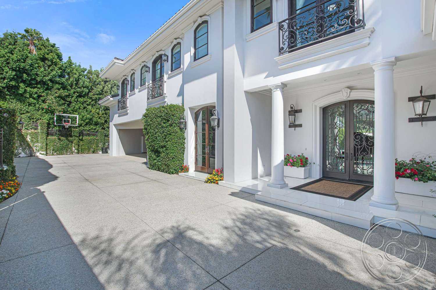 Medit 170 - exterior, white stucco walls, lush greenery, basketball hoop, landscaped flower beds, paved entryway, black wrought iron balcony railings, square columns, aggregate concrete driveway, double front doors with decorative glass