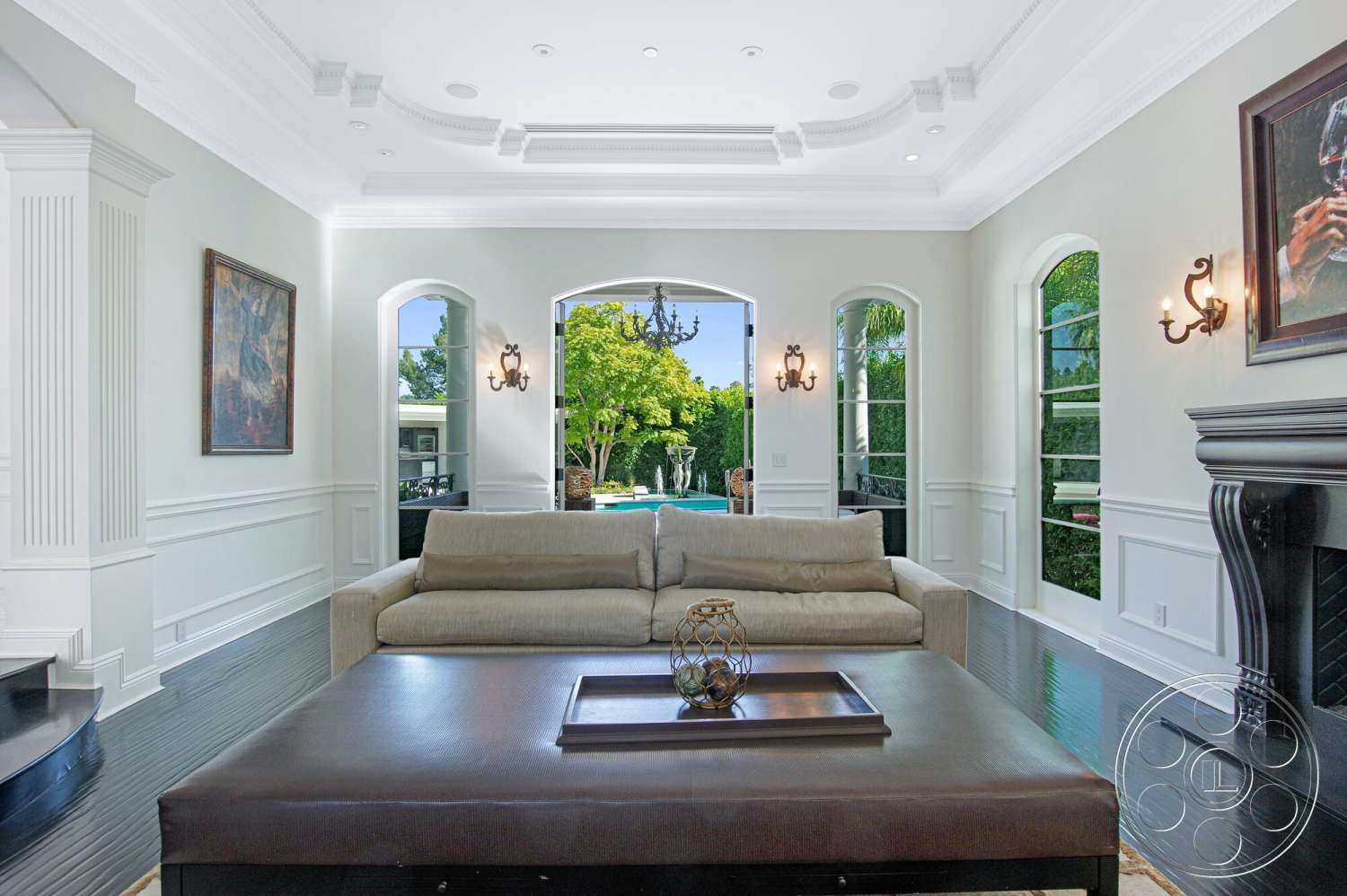 Medit 170 - artwork on wall, dark hardwood flooring, natural light from large windows, arched windows, white painted walls, contemporary sectional sofa, elegant chandelier lighting, traditional fireplace surround, vaulted ceiling with crown molding, open living room layout, wainscoting paneling, glass door leading to pool area, decorative table with geometric design, view of landscaped outdoor area