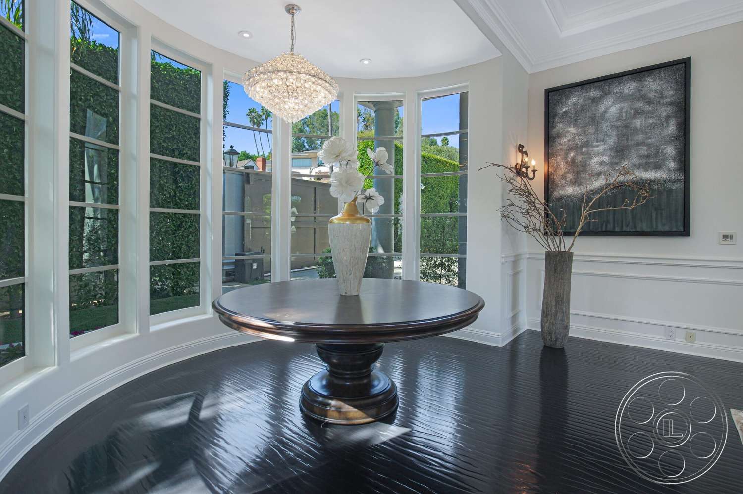 Medit 170 - high ceilings, floor-to-ceiling windows, round dining table, black hardwood flooring, white painted walls, natural light flooding in, curved window design, modern wall art, white crown molding, dark wood table surface, large crystal chandelier, minimalist floral arrangement