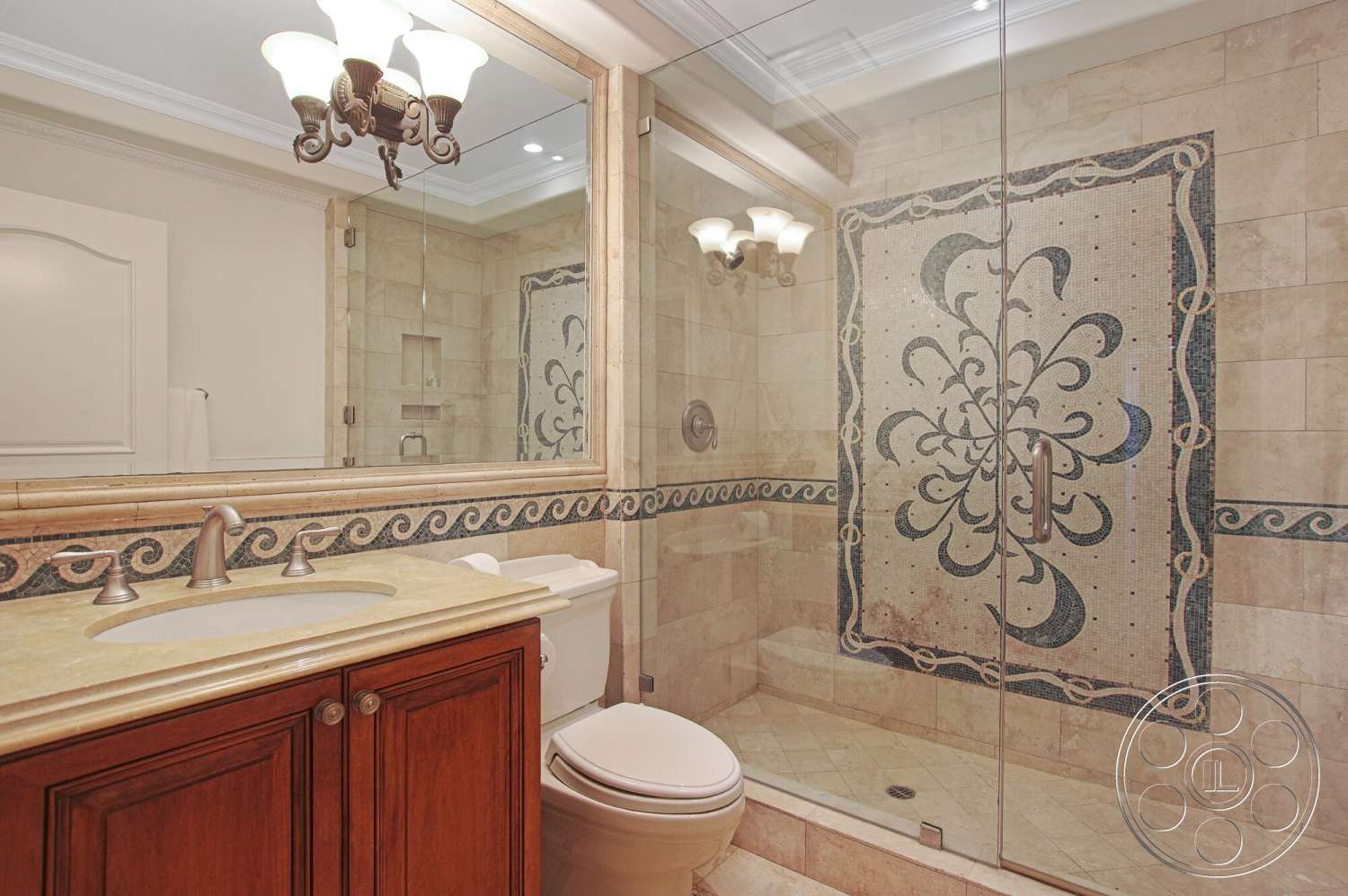 Medit 170 - glass shower enclosure, natural stone shower walls, brushed nickel faucets, white crown molding details, framed mirror with ornate details, travertine tile bathroom flooring, dark wood vanity with bronze fixtures, decorative tile backsplash with floral pattern, beveled edges on countertop, traditional style bathroom lighting