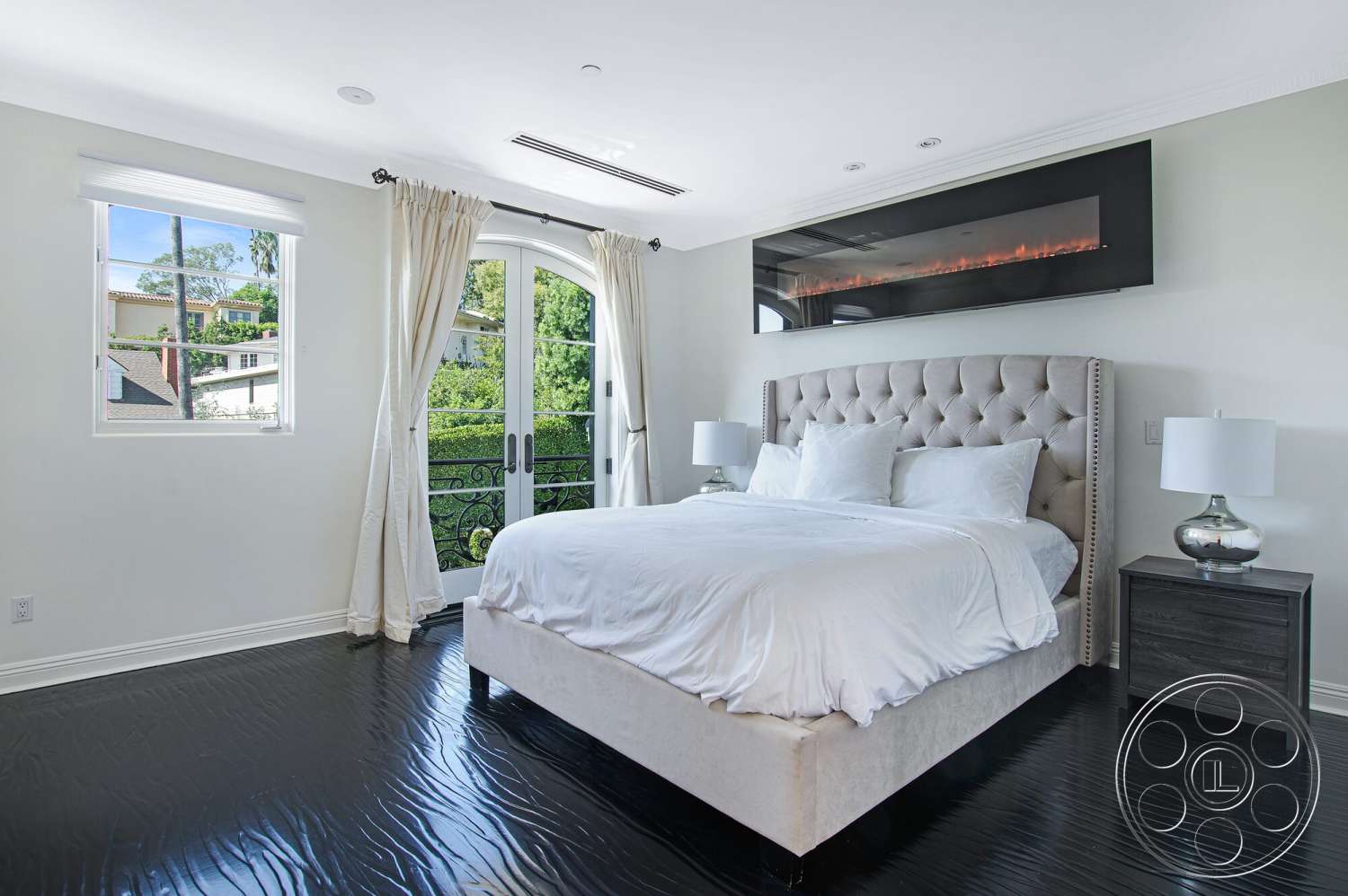 Medit 170 - floor-to-ceiling windows, white bedding, hardwood bedroom floors, neutral painted walls, glass balcony doors, dark wood nightstands, tufted upholstered bed frame, modern wall-mounted electric fireplace, decorative curtain panels, trimmed window casings, suburban view from window, chrome table lamps